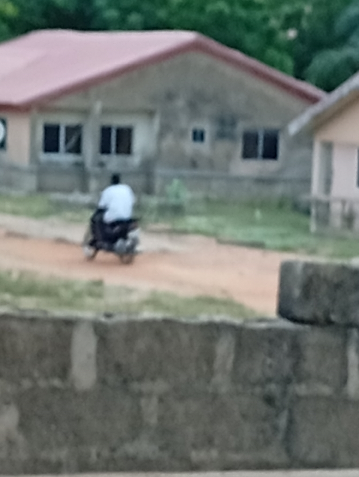 The image shows a man on a motorcycle on a dirt road in Adankolo, Nigeria. The man is wearing a white shirt. In the background are one-story buildings with red roofs. A block wall is in the foreground. The photo appears to be taken from behind the wall. The environment is suburban with some trees visible in the background.