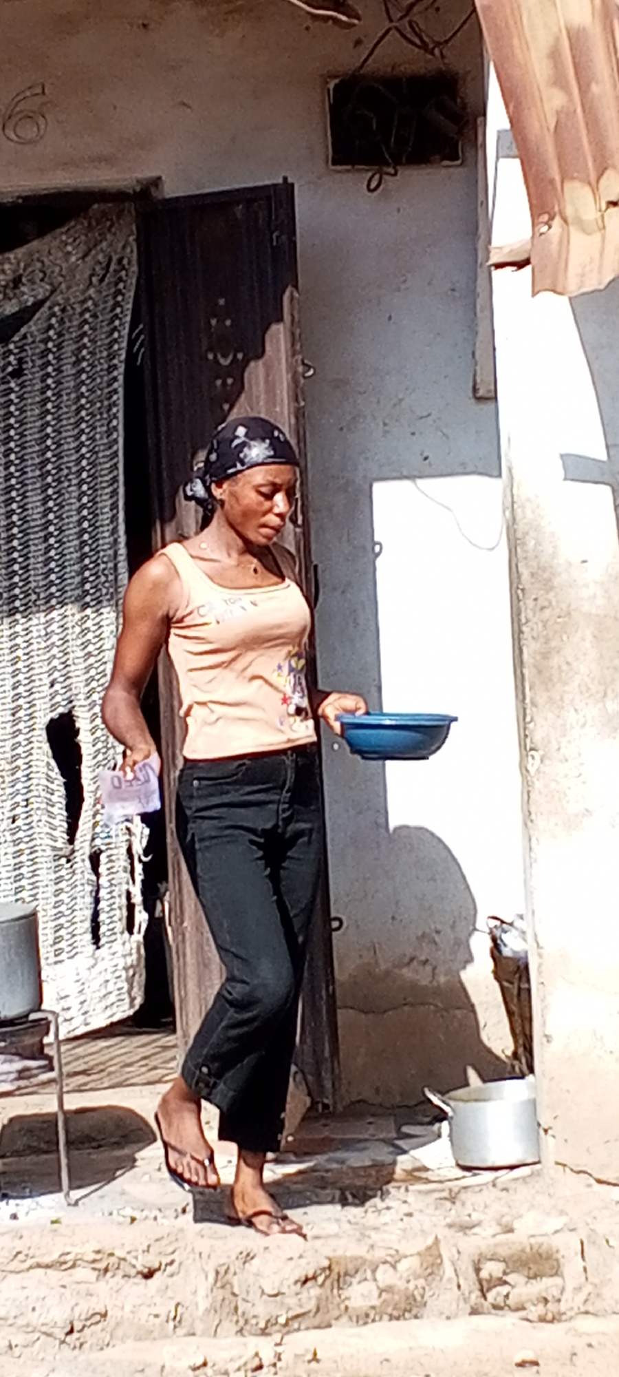 A woman stands in the doorway of a building in Sabon Tasha, Nigeria. She wears a tank top, black jeans, and flip-flops, and a headscarf. She holds a blue bowl in her right hand and folded bills in her left. The building has a dark wooden door and white walls, and there is a wicker screen to the left of the doorway. A metal pot is visible on the ground near the entrance. A small, barred window is visible above the door. The number six is faintly visible on the wall. The ground is uneven and appears to be made of stone or concrete.