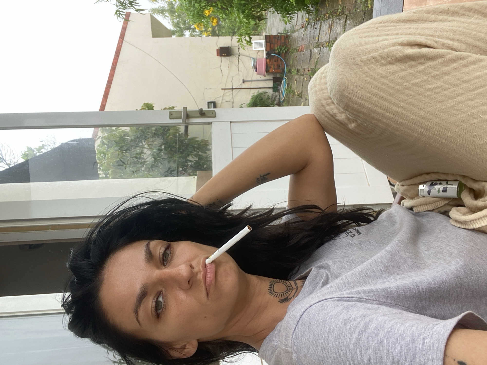 A woman with dark hair and light-colored eyes is depicted in a close-up, horizontally oriented shot. She is lying or reclining, looking towards the viewer with a neutral, relaxed expression, an unlit cigarette held between her lips. She wears a grey t-shirt, possibly with unreadable dark text near the neckline, and light beige or cream-colored, textured pants. A sun-like tattoo is visible on her neck, and another faint tattoo is on her upper left arm. Her right arm is bent and resting above her head. A white lighter with a floral design rests near her right hip on a folded piece of beige fabric or blanket.
The scene appears to be taken from indoors, looking out through a glass door or window with a white frame and a horizontal bar. The background reveals an outdoor setting, likely a backyard or side yard in a residential area of Mũi Né, Vietnam. The ground outside is visibly wet, indicating recent rain. A light-colored building with some cracks on its facade and a distinctive roofline is in the mid-ground. Various green plants, trees with yellow flowers, a brick structure, and plumbing pipes/hoses are also visible. The overall lighting suggests it is daytime, possibly an overcast day or immediately after rain.