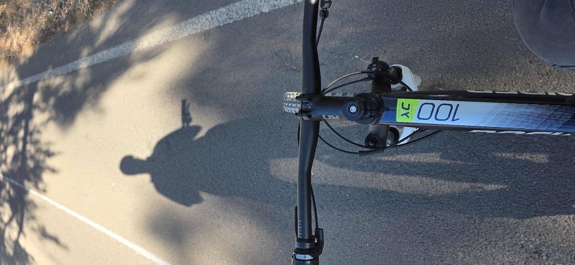The image shows an overhead view of a bicycle on an asphalt surface, likely a road or bike path. The bicycle's handlebars, front wheel, and a portion of the frame are visible. The frame has "XC 100" printed on it. The shadow of a person holding a camera is cast on the pavement, indicating they are taking the picture. The environment includes a white line on the asphalt and a shadow of trees or other vegetation along the edge of the road. The location is likely a recreational area, potentially a bike path along the coast near the city of Canet d'en Berenguer in Spain.