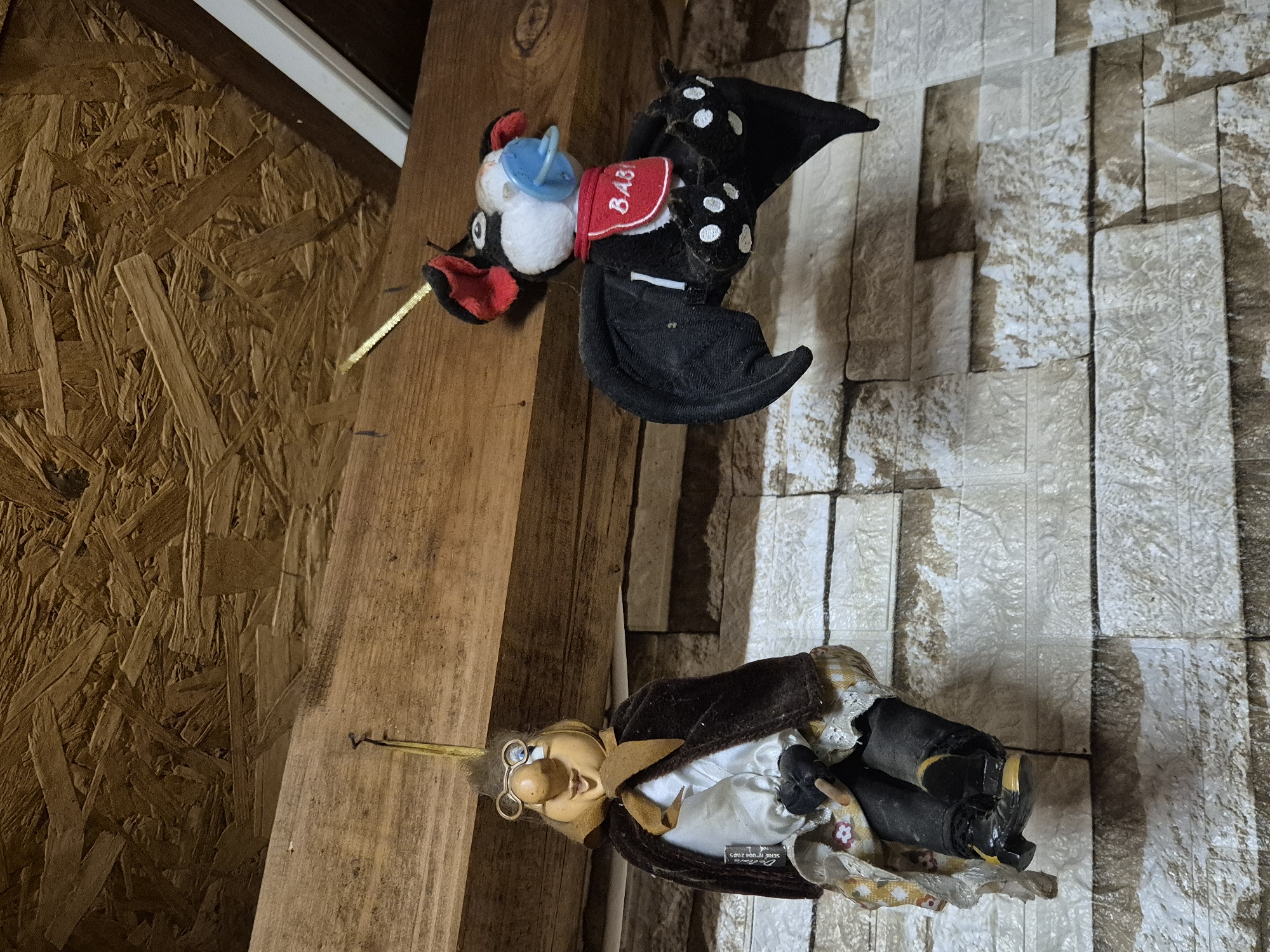 Two handmade dolls hang from a wooden beam attached to a ceiling made of oriented strand board. The doll on the left is a caricatured figure with large glasses, a prominent nose, and a yellow scarf, wearing a brown cape over a white shirt and a patterned skirt. The doll on the right is a plush bat with red ears, white markings on its wings and paws, a blue pacifier in its mouth, and a red bib that says "BABY". Both dolls are suspended by yellow strings. The wall behind the dolls is textured and appears to be made of faux stone or brick panels. The lighting suggests an indoor environment, possibly a workshop, garage, or unfinished room.