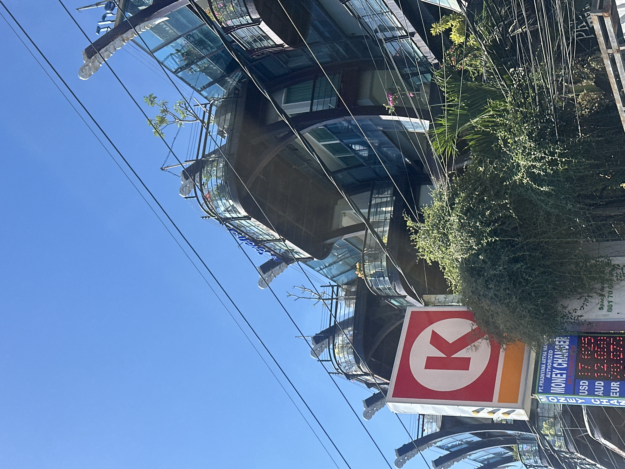 The image shows a modern building in Kuta, Indonesia, on a clear day with a bright blue sky. The building features curved balconies with glass railings and decorative white sculptures on its exterior. A prominent "K" logo within a red circle, indicating a Circle K convenience store, is visible on a sign on the left side of the frame. Below the "K" logo, a digital display shows currency exchange rates for USD, AUD, and EUR, identifying the establishment as a money changer. Lush green vegetation, including leafy bushes and palm fronds, partially obscures the lower part of the building and the signs. Multiple electrical wires crisscross the scene, suggesting an urban environment. The architecture and signage indicate a commercial area likely catering to tourists or local commerce.