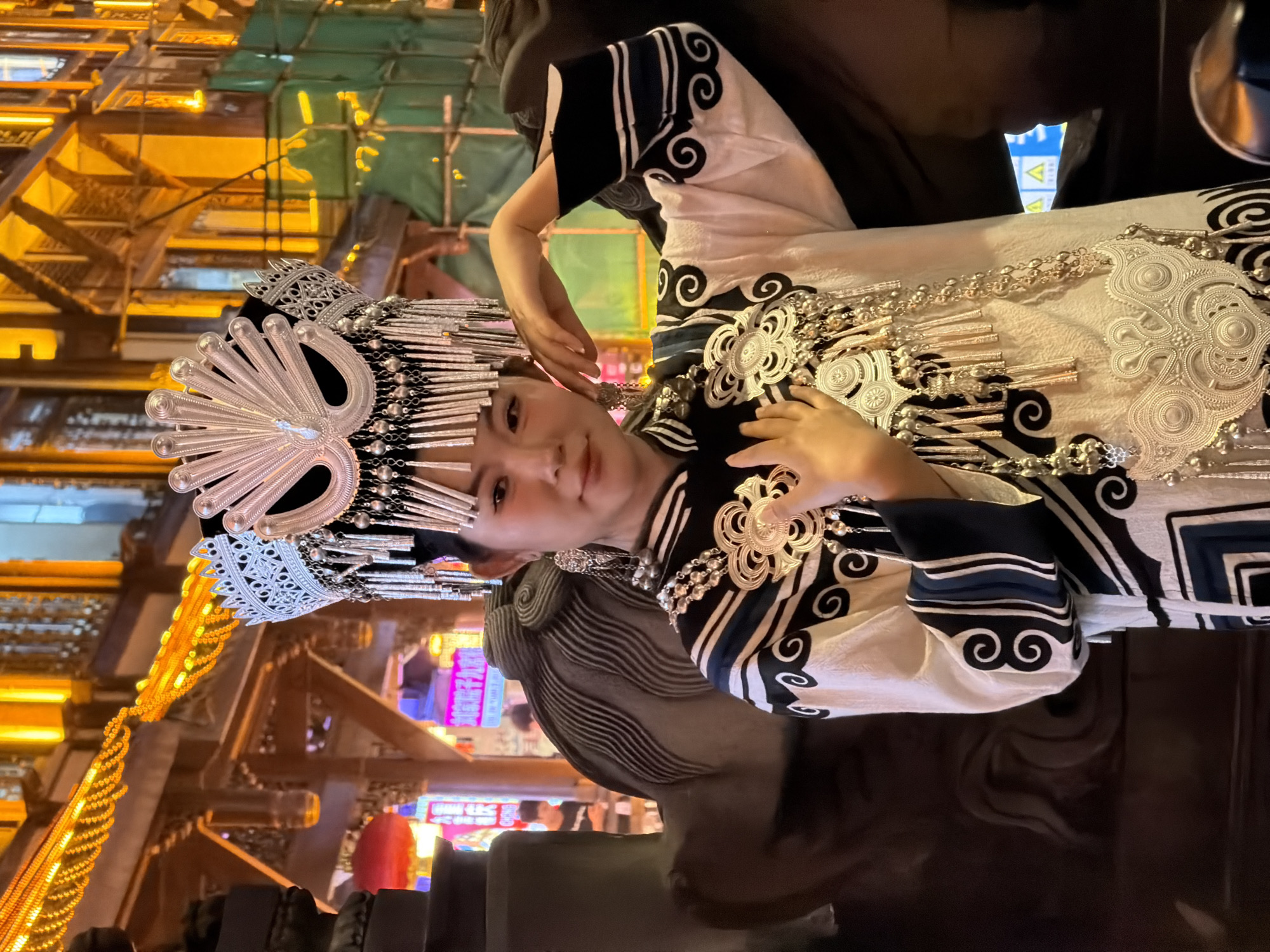 A young Asian woman stands in front of a dimly lit, ornate building in Yuzhong District, China. She wears a traditional Miao ethnic costume featuring a headdress adorned with silver filigree and intricate dangling ornaments, a multi-layered silver necklace, and a white tunic with black and blue geometric patterns. Her left hand is raised to her face, and her right arm is bent. The background shows traditional Chinese architecture with illuminated wooden eaves and colorful neon signs, suggesting a bustling night market or tourist area. The overall lighting is warm, with prominent yellow and orange hues from the building's illumination and some green scaffolding visible on the right.