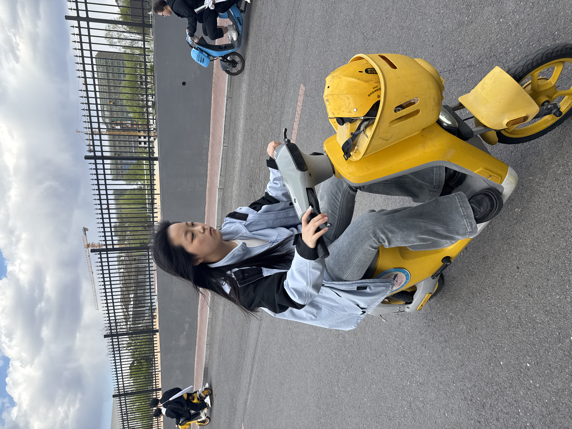 The image, rotated 90 degrees counter-clockwise, depicts a daytime outdoor scene. A young Asian woman with long dark hair is seated on a bright yellow electric scooter. She wears a light blue and black jacket over a white top, paired with grey jeans and black shoes. Her hands are on the scooter's grey handle or control mechanism. A yellow helmet, possibly belonging to the scooter, rests on the front part of the vehicle.
In the background, another person can be seen on a blue electric scooter. The setting is a paved area, likely a street or pathway, adjacent to a tall black metal fence. Beyond the fence, a construction site is visible, featuring a large building under construction covered in scaffolding and a prominent yellow crane. The sky above is partly cloudy. The overall environment suggests an urban or developing area in Zhongtun, China.
Notable details include Chinese characters on the yellow helmet, which likely include a safety instruction such as "请正确佩戴安全帽" (Please wear the helmet correctly). A circular sticker on the side of the yellow scooter contains faint text and the numbers "1951", possibly part of a brand logo or historical marker. The woman's expression is neutral and calm.