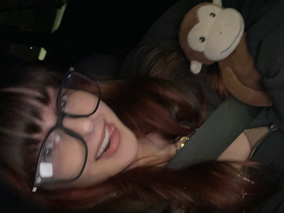 A female individual is seated inside a vehicle in Raanana, Israel, under low-light conditions, likely at night. She has dark hair with reddish undertones and bangs, and wears large black-framed glasses. Her mouth is slightly open, with her tongue partially extended in a playful expression. A golden chain necklace is visible around her neck, and a dark seatbelt crosses her chest diagonally. Resting on her right side, partially against her dark zippered garment, is a brown and tan stuffed monkey toy featuring a light-colored face and a stitched smile.