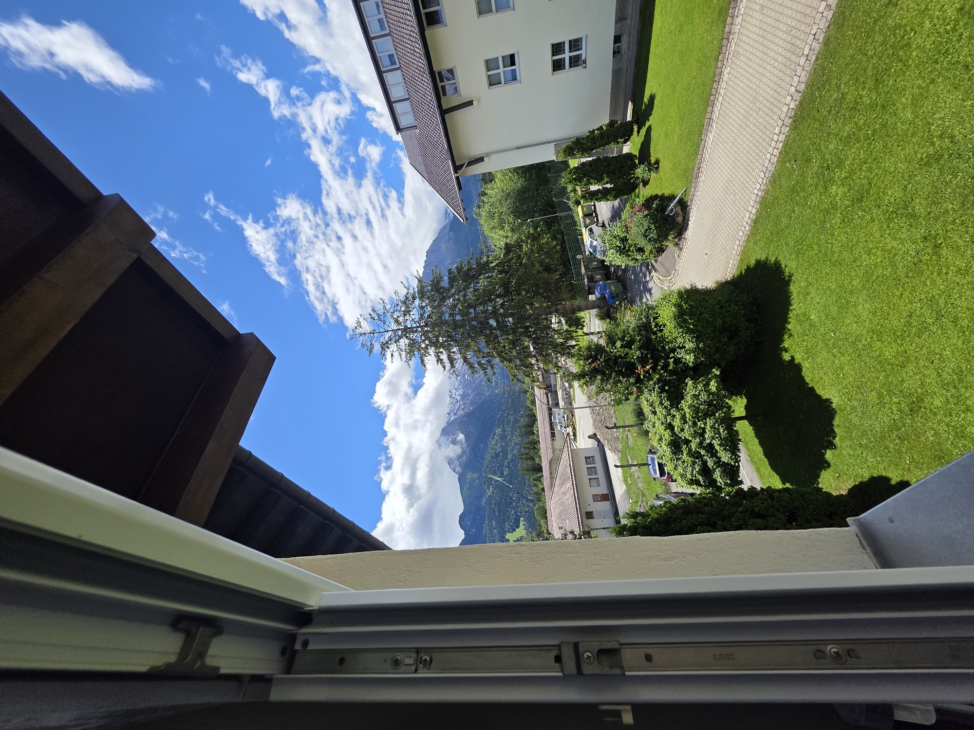 This is an outdoor, eye-level, medium shot of a mountain landscape as seen from a window. The foreground is dominated by the open window frame on the left and a section of a building on the right. The window frame is grey metal and cream-colored. The building is white with a brown, tiled roof and several windows. Beyond the immediate structures, a well-maintained lawn is visible, with a paved walkway curving through it. In the middle ground, there are several trees, including a tall evergreen and a smaller, bushier tree casting a shadow on the grass. A road with a few parked cars and some trash receptacles is also present. The background features a majestic mountain range with snow-capped peaks, partially obscured by clouds. The sky is a vibrant blue with scattered white clouds. The scene appears to be set in a residential or institutional area, possibly near a mountainous region.