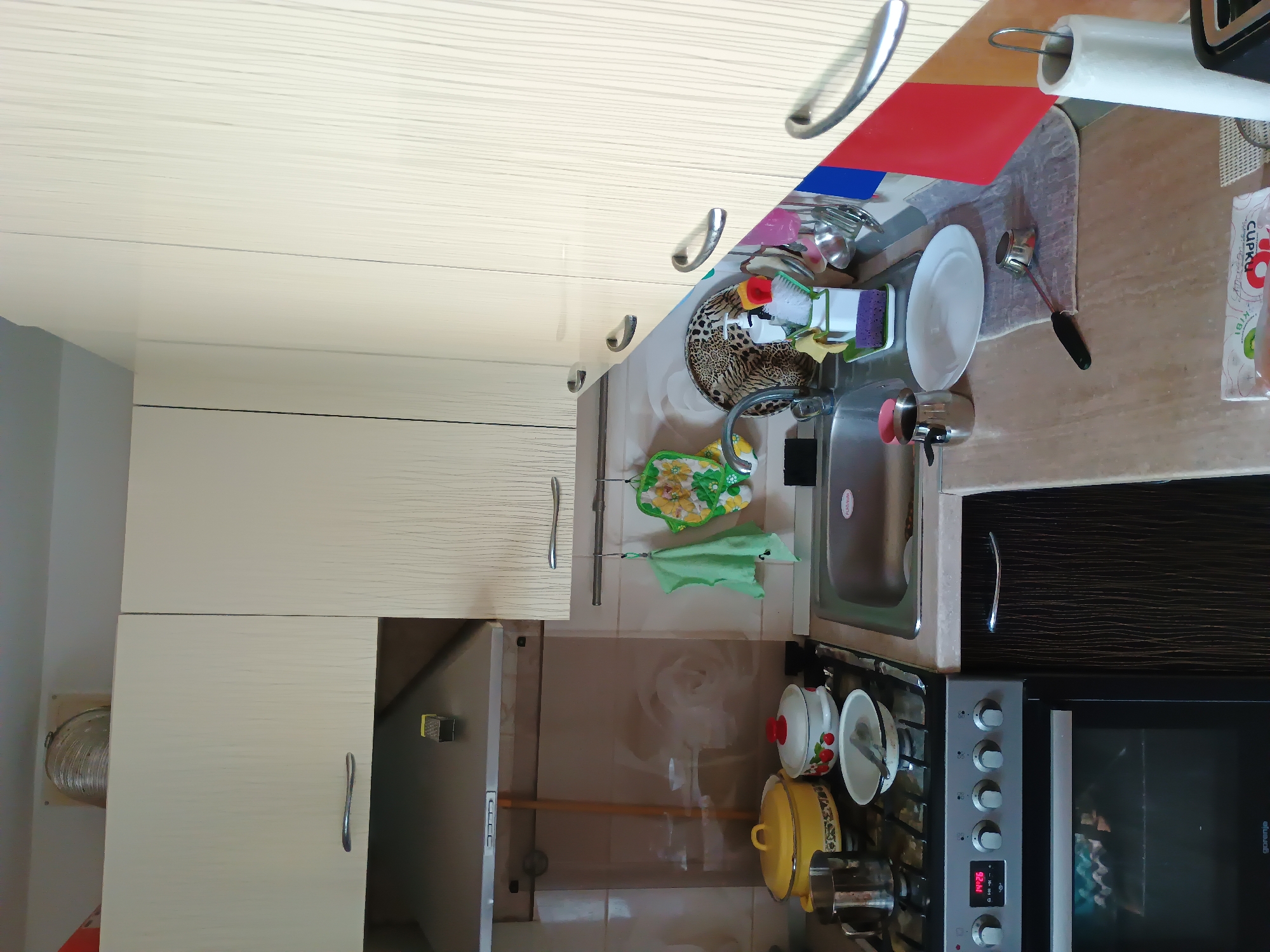 The image depicts a domestic kitchen scene. The kitchen features light-colored cabinets with a textured wood grain finish, a stainless steel sink, and a gas stove with an oven. On the stove, there are several pots, including a yellow one and a white one with red fruit detailing. A metal pitcher is also present. Above the stove, a stainless steel range hood is installed. Next to the sink, there is a dish rack holding utensils and cleaning supplies. Two green and yellow patterned oven mitts or pot holders are hanging from a rack above the sink. The countertop is a light beige material with a speckled pattern. The overall environment appears to be a functional, everyday kitchen space. There is no indication of the specific location within the city of Zaporizhzhia, Ukraine.
