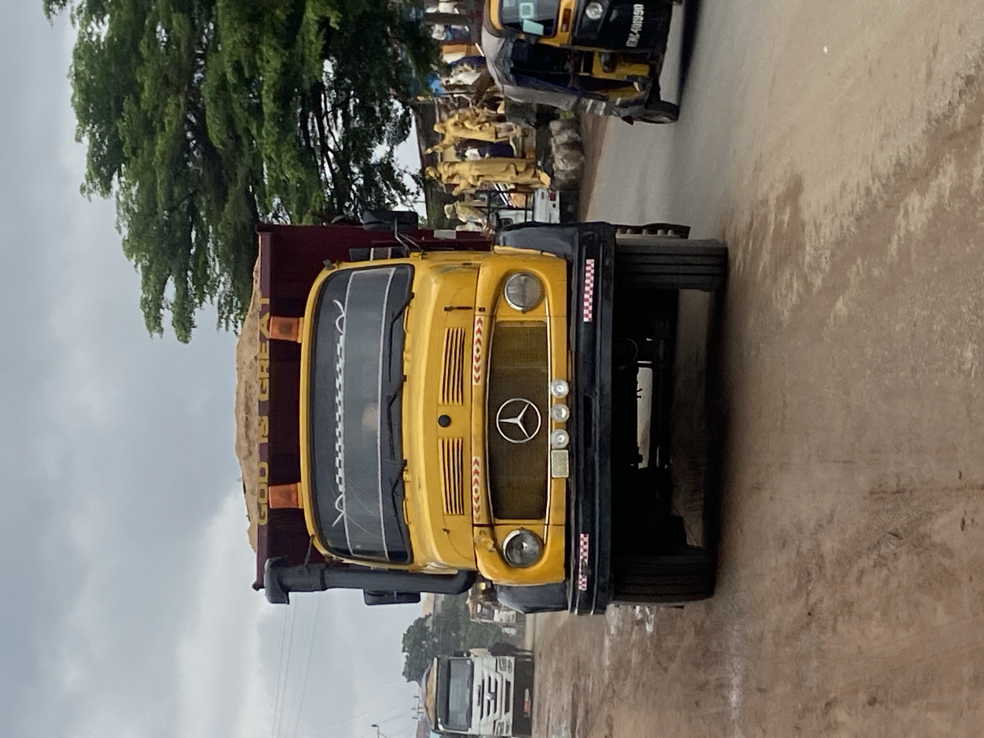 In Nkpor, Nigeria, a bright yellow Mercedes-Benz dump truck, loaded with sand, is the primary subject in the foreground, facing the camera. The truck has a dark red cargo bed and an orange flashing light on top. The Mercedes-Benz emblem is visible on the grille. Behind the truck, to the right, a yellow and black auto-rickshaw is partially visible. Further in the background, a white truck is seen on the left side of the road. The environment appears to be a dusty road with some trees and buildings in the distance. The sky is overcast. In the mid-ground, to the right of the yellow truck, a group of golden statues are displayed.