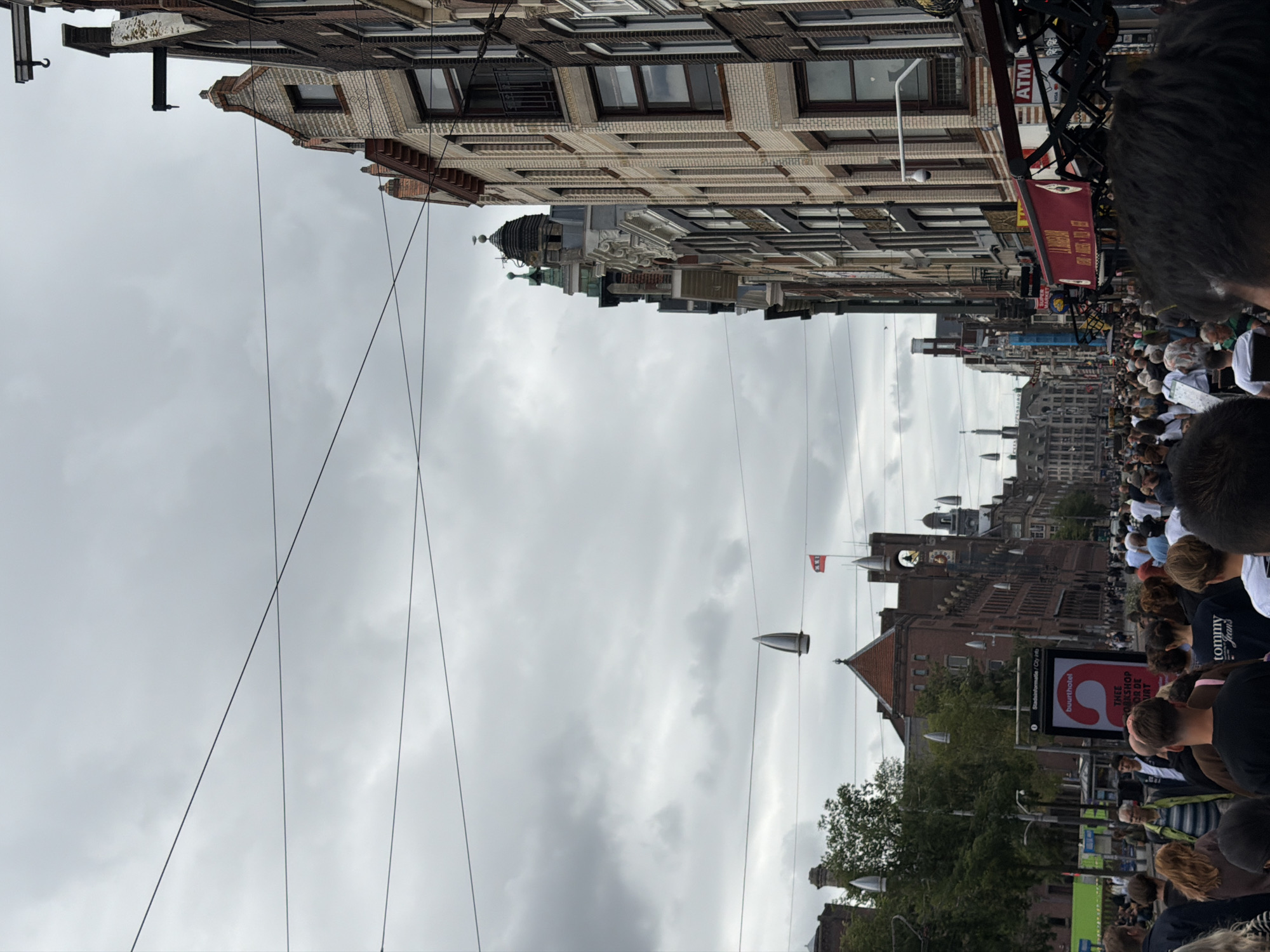 The image depicts a crowded street scene in Amsterdam, Netherlands, under an overcast sky. The street is lined with traditional Dutch canal houses, characterized by their tall, narrow facades and distinctive gables. Many of these buildings appear to be shops or businesses, with awnings and signs visible, including an ATM. A large digital billboard for "buurthotel" with a prominent "A" graphic is situated on the left side of the street.
The street is filled with a dense crowd of people, many walking in both directions. They are dressed in casual clothing, suggesting it is a public space for leisure or tourism. Some individuals are looking towards the buildings, while others are engaged in conversation. The atmosphere appears lively due to the large number of people.
Overhead, a network of wires crisscrosses the sky, typical of urban infrastructure in older European cities. Several decorative, pointed objects are suspended from these wires, adding a unique visual element to the scene. A red and white flag is visible on a flagpole in the distance, atop one of the buildings. The overall impression is a busy, urban environment in Amsterdam, likely a popular area for pedestrians and commerce.