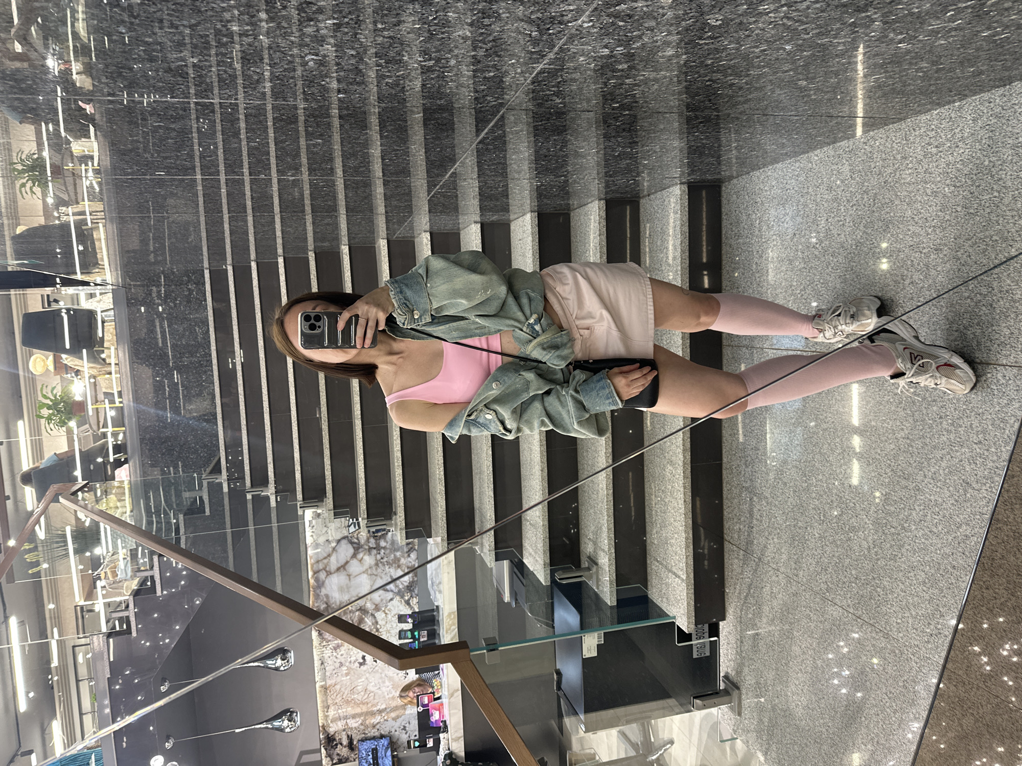 A person is taking a mirror selfie on a wide, modern staircase. The staircase features dark, polished granite steps with silver trim, interspersed with black marble risers. A glass railing runs along the side of the stairs. The person is wearing a pink tank top, a light blue denim jacket, light pink shorts, and tall light pink socks with white athletic shoes. They are holding a smartphone to take the photo. The reflection in the mirrored wall behind them shows the interior of a building, possibly a shopping mall or office building, with more staircases, planters with greenery, and lighting fixtures visible. The scene appears to be indoors, likely during the daytime, as indicated by the lighting. There is no visible text in the image.