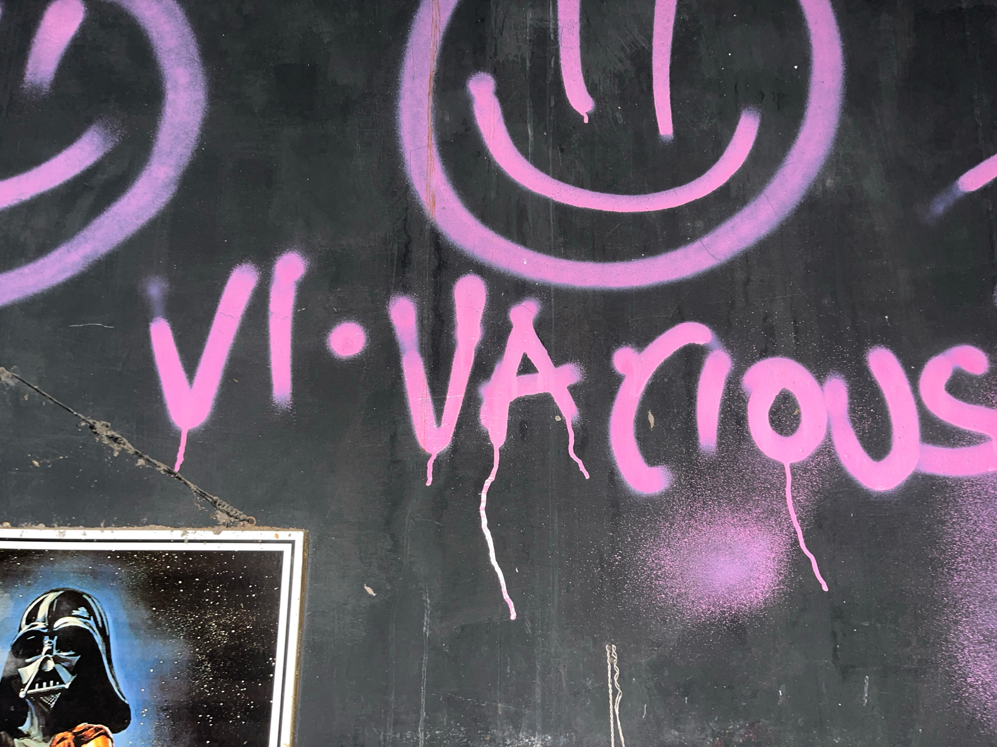 A close-up, eye-level shot captures a black, textured wall adorned with graffiti. In the upper half of the image, two large, pink spray-painted smiley faces are visible, their curved lines subtly highlighted by a lighter shade of pink. Below the smiley faces, a line of pink graffiti spells out "VICTIOUS," with a pink dot preceding the "V." The letters are unevenly applied, with some areas showing thicker paint and drips. Below the graffiti, a framed poster featuring Darth Vader is partially visible in the lower left corner, his iconic helmet and menacing expression rendered in dark tones against a starry blue background. The wall appears weathered, with some cracks and dirt marks contributing to its rough texture. The scene is set outdoors, likely on a street in Surakarta, Indonesia.