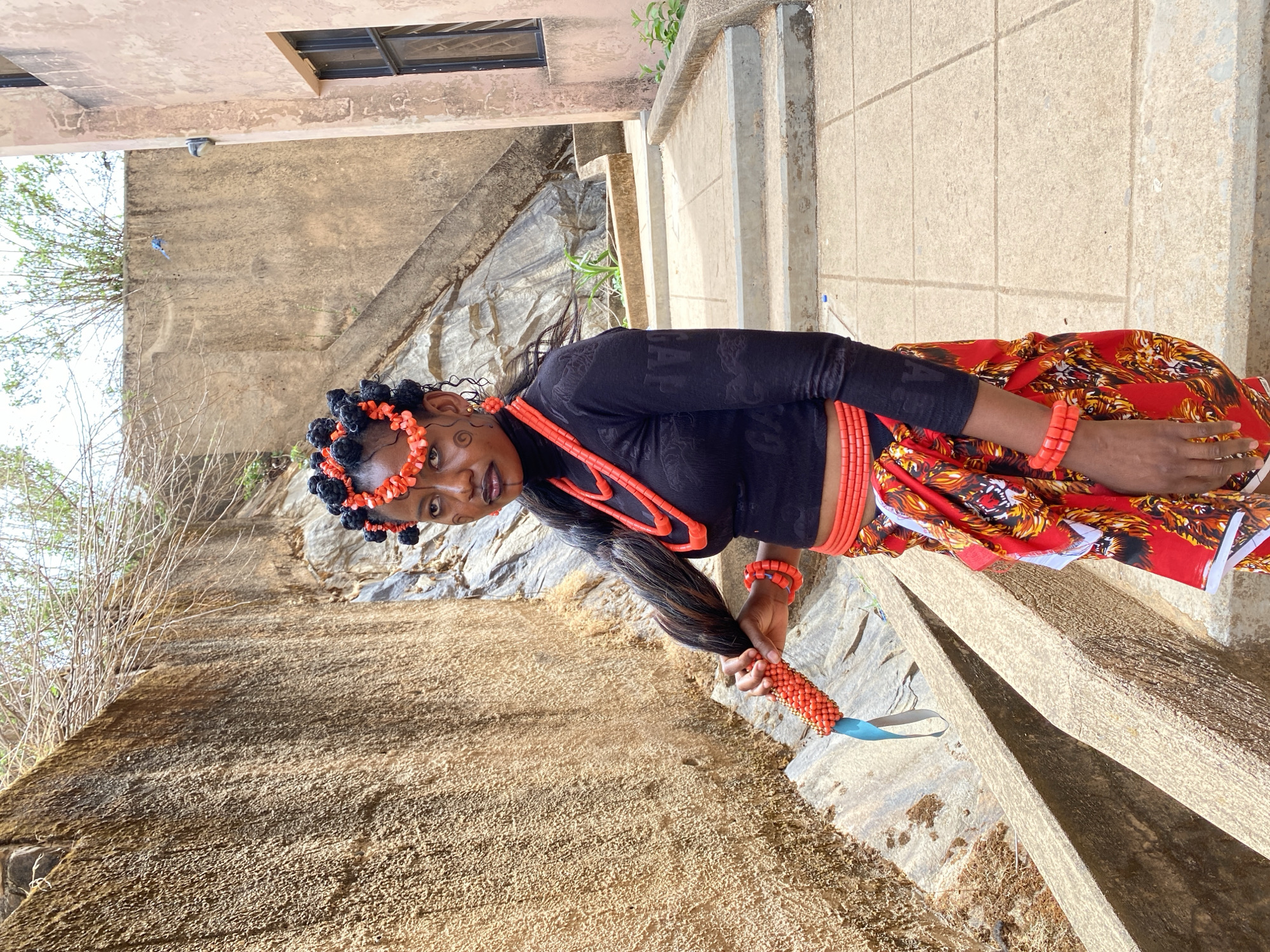 A medium shot, taken outdoors, shows a woman in Gwong, Nigeria, standing on a stone structure. The structure appears to be a wall or retaining structure on a hillside. The woman is wearing traditional garb, including a black long-sleeved shirt with patterns, a red and orange patterned skirt, and orange beaded jewelry including a headpiece, necklaces, and bracelets. Her hair is styled in braids with black beads. She is holding a beaded object with a blue loop in her right hand. The background is comprised of a stone wall with some vegetation. The light is bright and appears to be daylight.