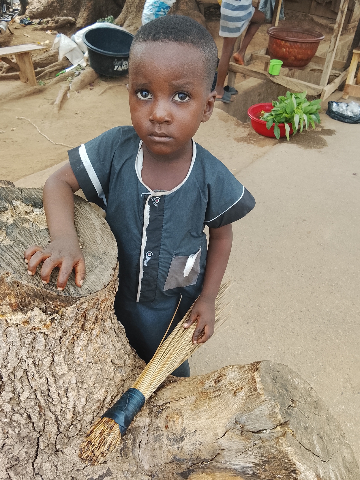 The image captures a young child standing outdoors in what appears to be a semi-urban or rural setting in Akwanga, Nigeria. The child, likely a boy, has dark skin, short dark hair, and large, expressive dark eyes that look directly at the viewer with a serious, captivating gaze. He is wearing a dark grey or blue traditional tunic with white trim along the collar and sleeves, and small white buttons down the front. A small, patched pocket is visible on the right side of his tunic.
He is leaning with his left hand on a large, rough-hewn wooden tree stump or log, which dominates the lower left portion of the frame. Resting on another piece of wood next to him is a traditional broom made of bundled straw or reeds, bound with a dark, possibly black, wrapping.
In the background, the scene reveals a dirt ground mixed with some paved areas, indicative of a street or market. Various items associated with daily life or commerce are visible: a black plastic basin with faint text "FAMILY" on it, a brown woven basket, a bright green plastic cup, and a red plastic bowl filled with fresh green leafy vegetables (possibly spinach or similar greens). Further back, partial legs of another person wearing striped shorts and sandals are visible, suggesting other people are present in the vicinity. The roots of a large tree are also evident in the background.
The lighting suggests it is daytime, with clear visibility. The child's expression is calm and still, adding a sense of quiet observation to the moment.