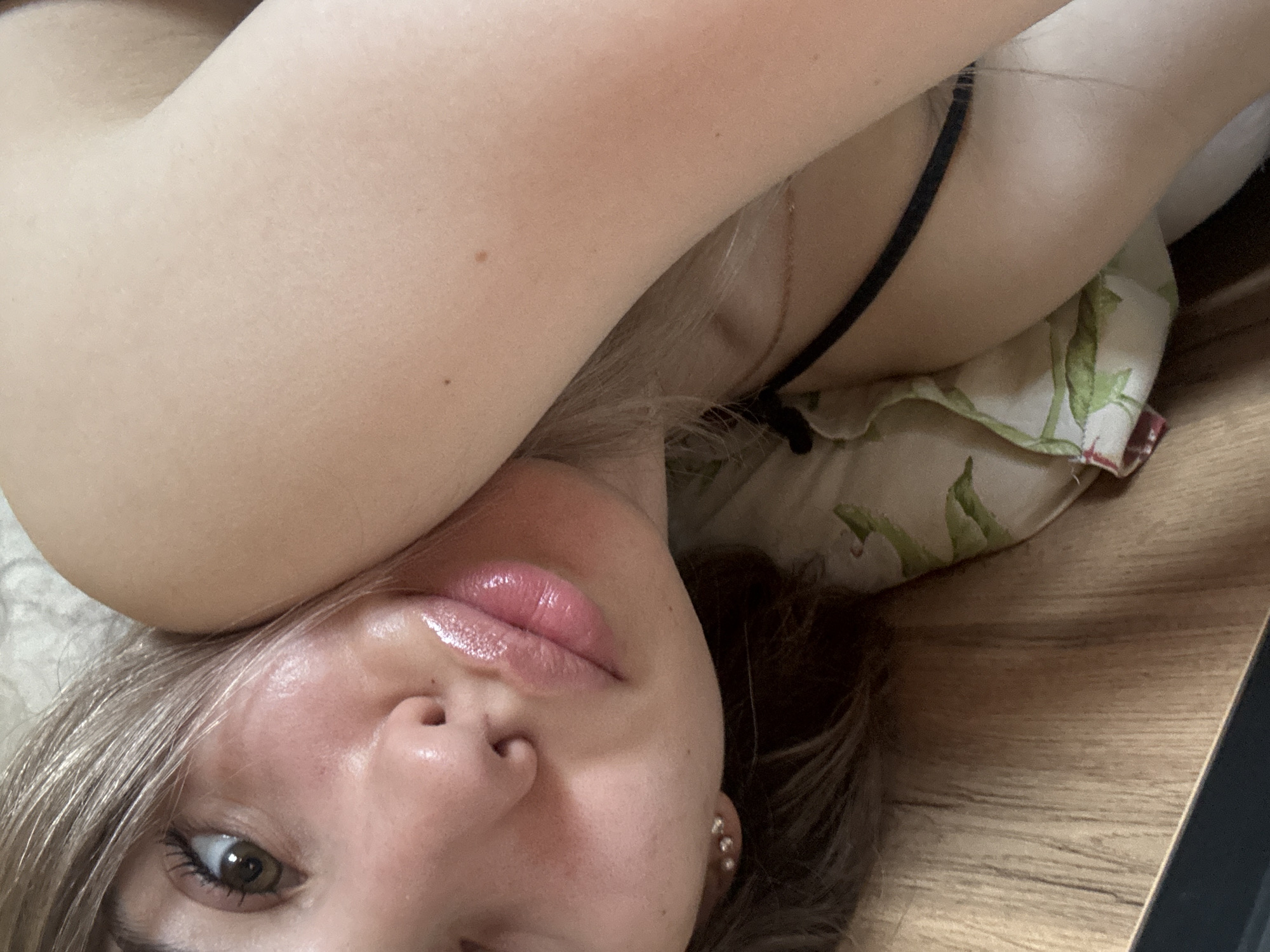 A close-up, slightly angled selfie of a fair-skinned individual with light brown hair and green eyes. The person is lying down, possibly on a pillow with a floral pattern. A thin, black strap is visible, possibly from a garment. The background includes a wooden surface. The location is not discernible.