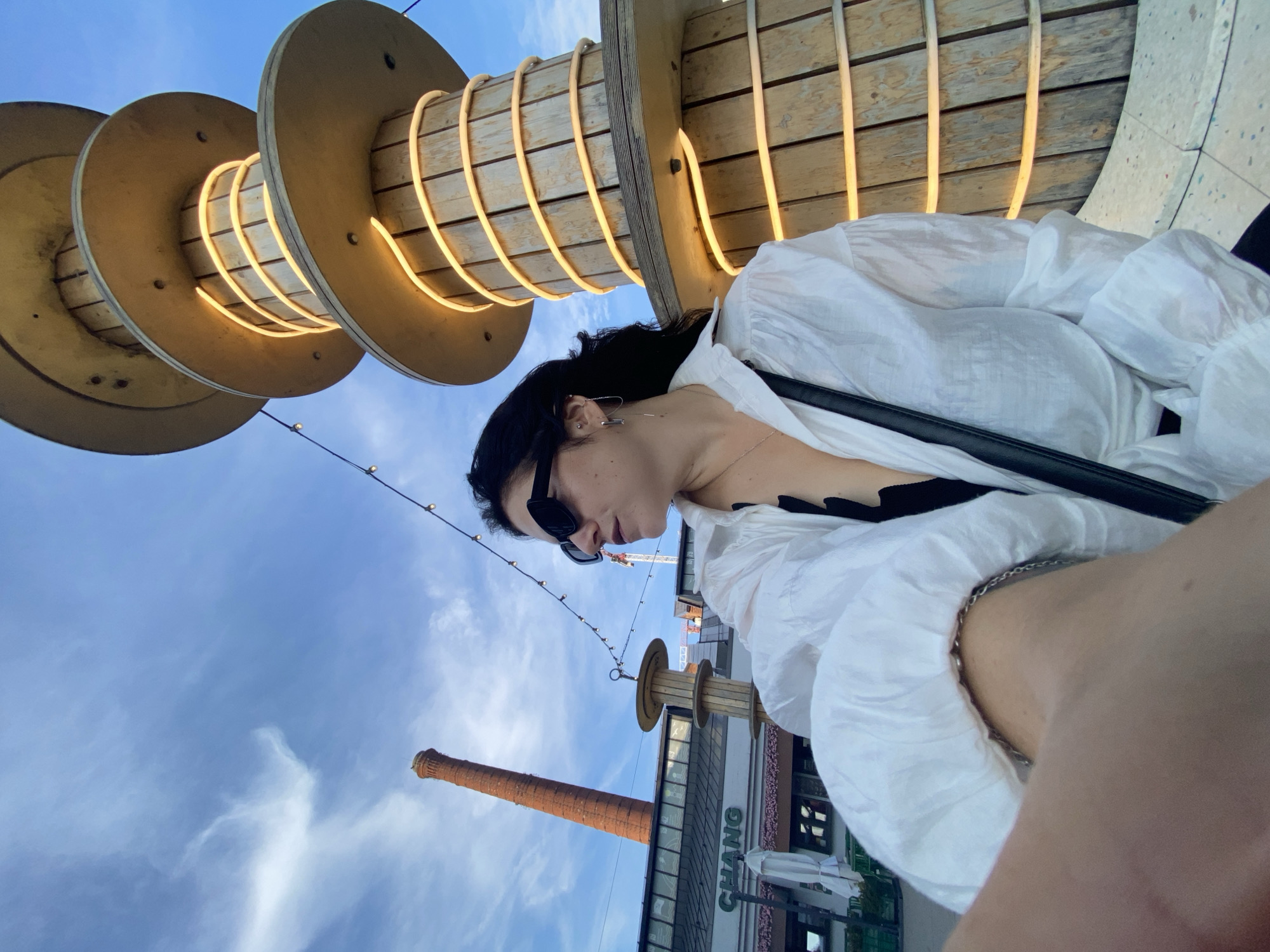 In Saint Petersburg, Russia, a young woman with dark hair and sunglasses is captured in a selfie against a backdrop of a partly cloudy blue sky. She is wearing a white collared shirt, open at the neck to reveal a black garment beneath, and a black crossbody bag. To her right, a tall, multi-tiered wooden structure is illuminated by warm, horizontal light strips. In the background, a red brick industrial chimney rises skyward, and string lights crisscross the upper portion of the frame. A building with the sign "CHANG" is visible to the left. The scene appears to be an outdoor public space, possibly a waterfront or entertainment area, given the presence of the industrial elements juxtaposed with modern design and lighting.