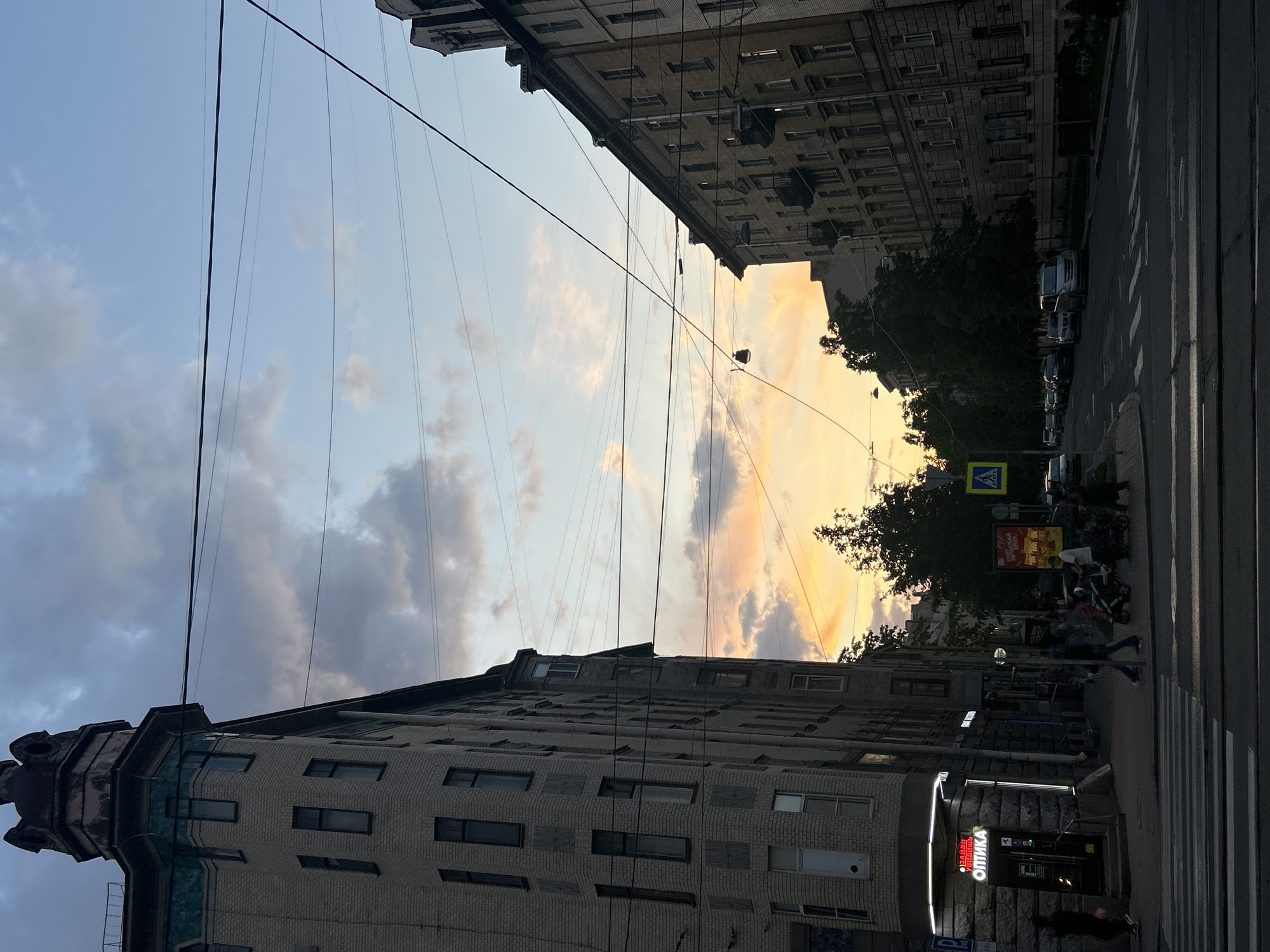 This image captures an urban street scene in Saint Petersburg, Russia, at what appears to be dusk or sunset. The perspective is looking upwards from street level, showing a narrow view of the sky between two tall, multi-story buildings that line the street.
The sky exhibits a gradient of colors, transitioning from a cooler blue with scattered clouds on the left to warm, vibrant orange, yellow, and pink hues on the right, indicative of a sunset. Numerous dark overhead power lines or trolley wires crisscross the sky between the buildings.
On the left side, a brick building with many windows extends upwards, featuring a distinct, ornate corner structure at its top. On the right side, another building, also brick, houses a ground-floor commercial establishment.
Below the buildings, a street is visible with distinct white zebra crossing markings. Several cars are parked along the right side of the street, and dark trees are interspersed between the building and the street. Pedestrians can be seen walking on the sidewalks and near the crosswalk. A blue and white pedestrian crossing sign is visible on the right.
Visible text includes a brightly lit sign on the right building that reads "ОПТИКА" (OPTICS) in Cyrillic, with smaller text below, possibly "ГЛАЗАМ УДОВЛЕТИШЬ" (EYES WILL BE SATISFIED). Further down the street on the right, a poster shows partial Cyrillic text, including "ТВОЙ ИДЕАЛЬНЫЙ..." (YOUR IDEAL...). The scene portrays a typical urban evening with a blend of historical architecture and modern city life.