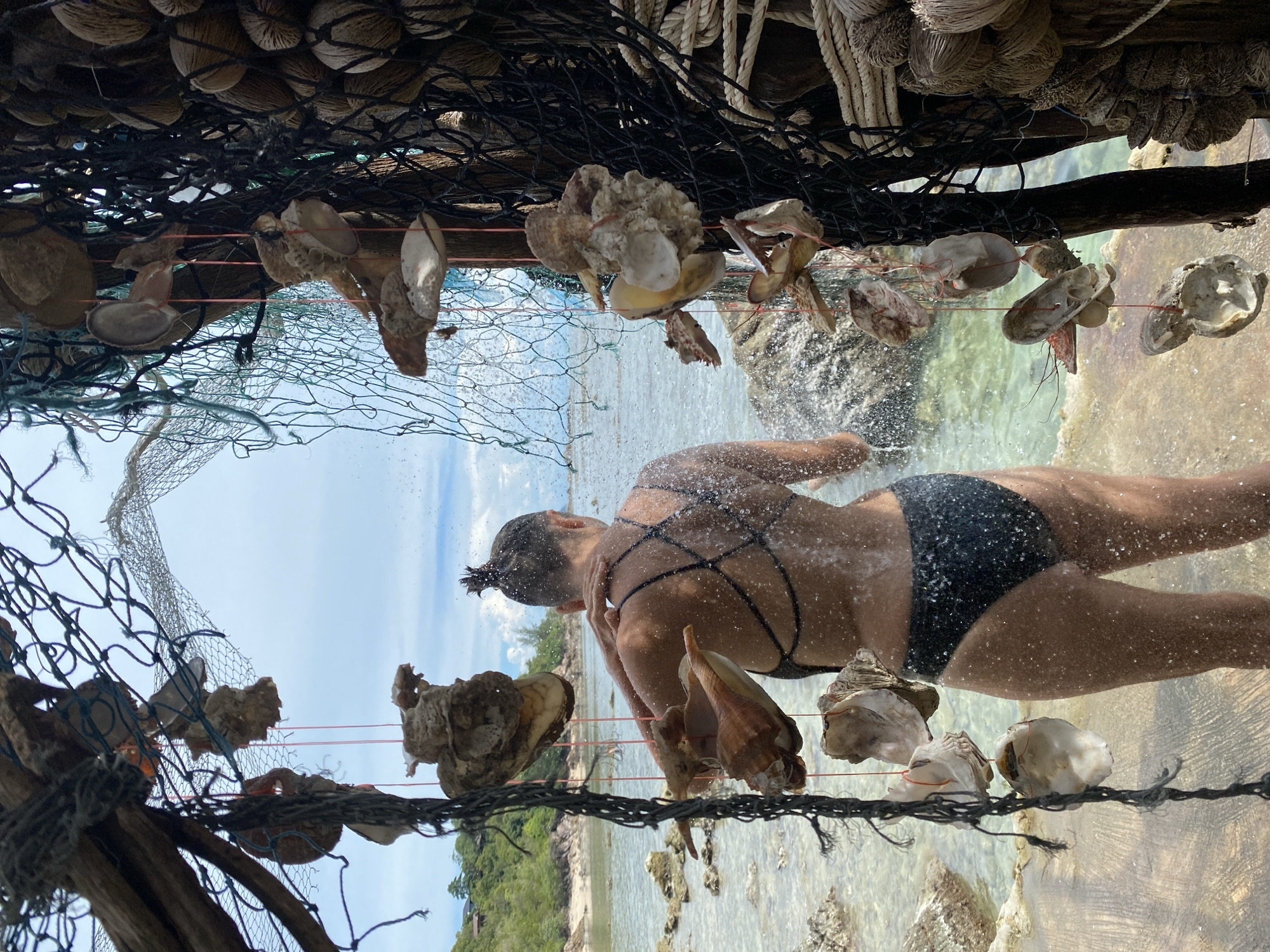 In ตำบลเกาะพะงัน, Thailand, a person in a black bikini stands with their back to the camera, facing a beach with clear, shallow water. They are holding their left hand to their shoulder, and water is spraying from the left side of the frame, indicating they are under a shower or being splashed. The scene is framed by a rustic structure made of driftwood and fishing nets, adorned with shells and dried seed pods. The background shows a lush, green hillside and a cloudy sky. The water is calm and reflects the sky, with some rocks visible in the shallows.