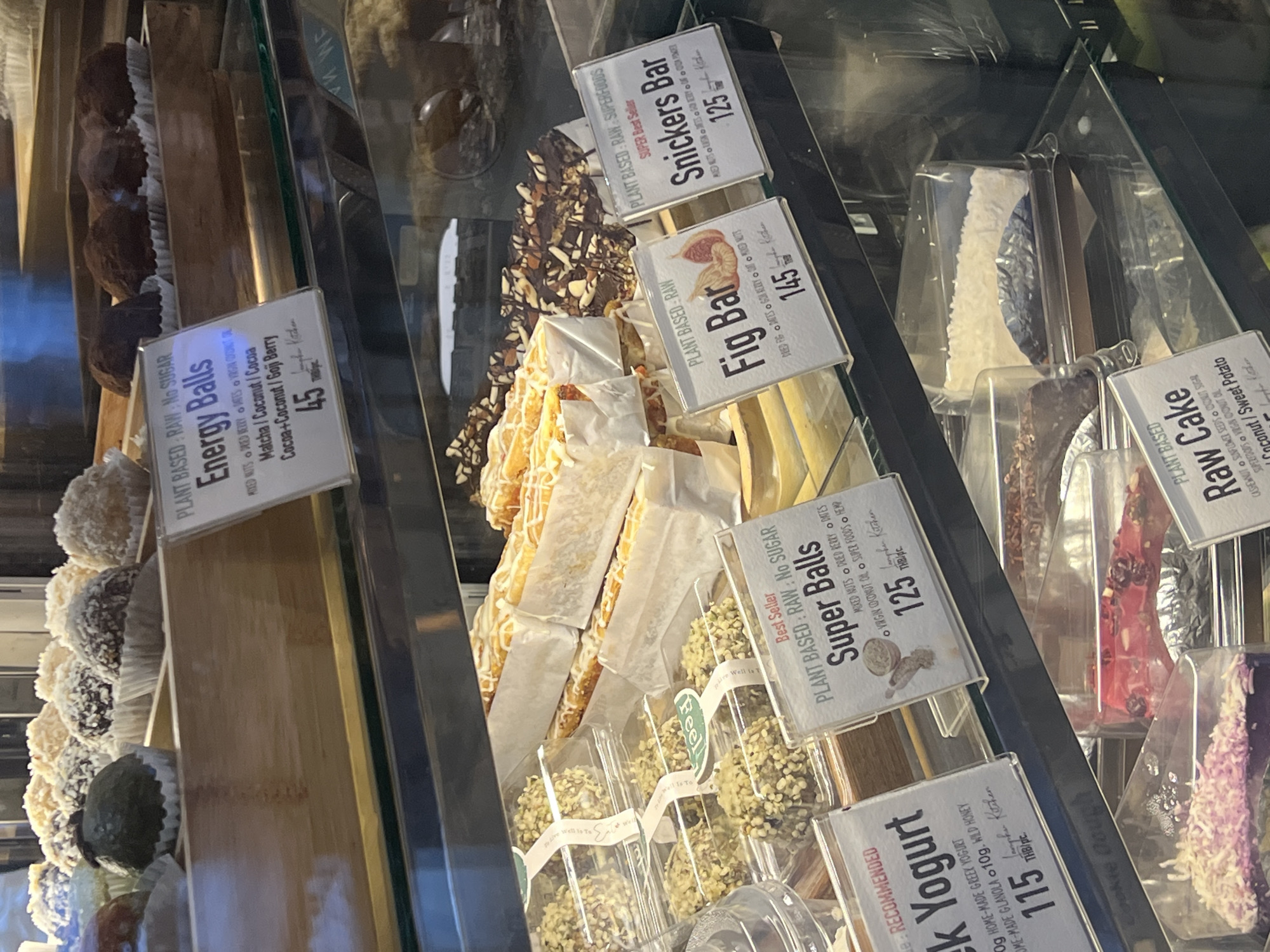 The image shows a display case at a bakery or health food store, likely in Ko Samui District, Thailand. The display case is filled with various desserts and snacks. In the top section, there are round, bite-sized treats, some coated in coconut flakes and others in chocolate. A sign in front of these reads "Energy Balls" and lists flavors like Matcha/Coconut/Cocoa and Cocoa+Coconut/Goji Berry, priced at 45 THB.
Below this, there are rectangular bars with white icing, and next to them, dark chocolate bars topped with nuts. Signs indicate "Fig Bar" priced at 145 THB and "Snickers Bar" (likely a healthier version) priced at 125 THB.
Further down, individual servings of what appear to be yogurt with granola are displayed in plastic containers, with a sign for "Greek Yogurt" at 115 THB. Other items visible include what might be a "Raw Cake" with coconut topping and possibly another cake-like item with pink and red fruit pieces. The overall environment is well-lit, showcasing the products for sale. The presence of Thai Baht (THB) on the price tags indicates the location as Thailand.