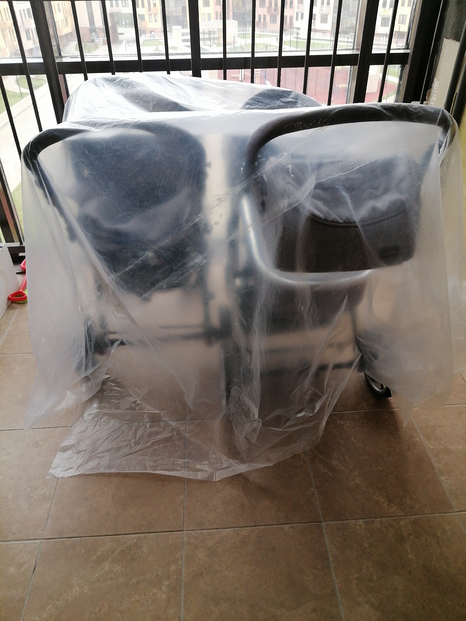 On a brown tiled floor, under a covered patio with black metal railing, is furniture wrapped in a transparent plastic sheet. The setting appears to be a covered balcony or patio area, with the railings providing a view of other buildings and green spaces in the background. The plastic sheet obscures details of the furniture beneath, with only the vague shapes of chairs visible. The scene is indoors, likely an apartment or similar residential space. A red plastic toy or object is partially visible near the side.