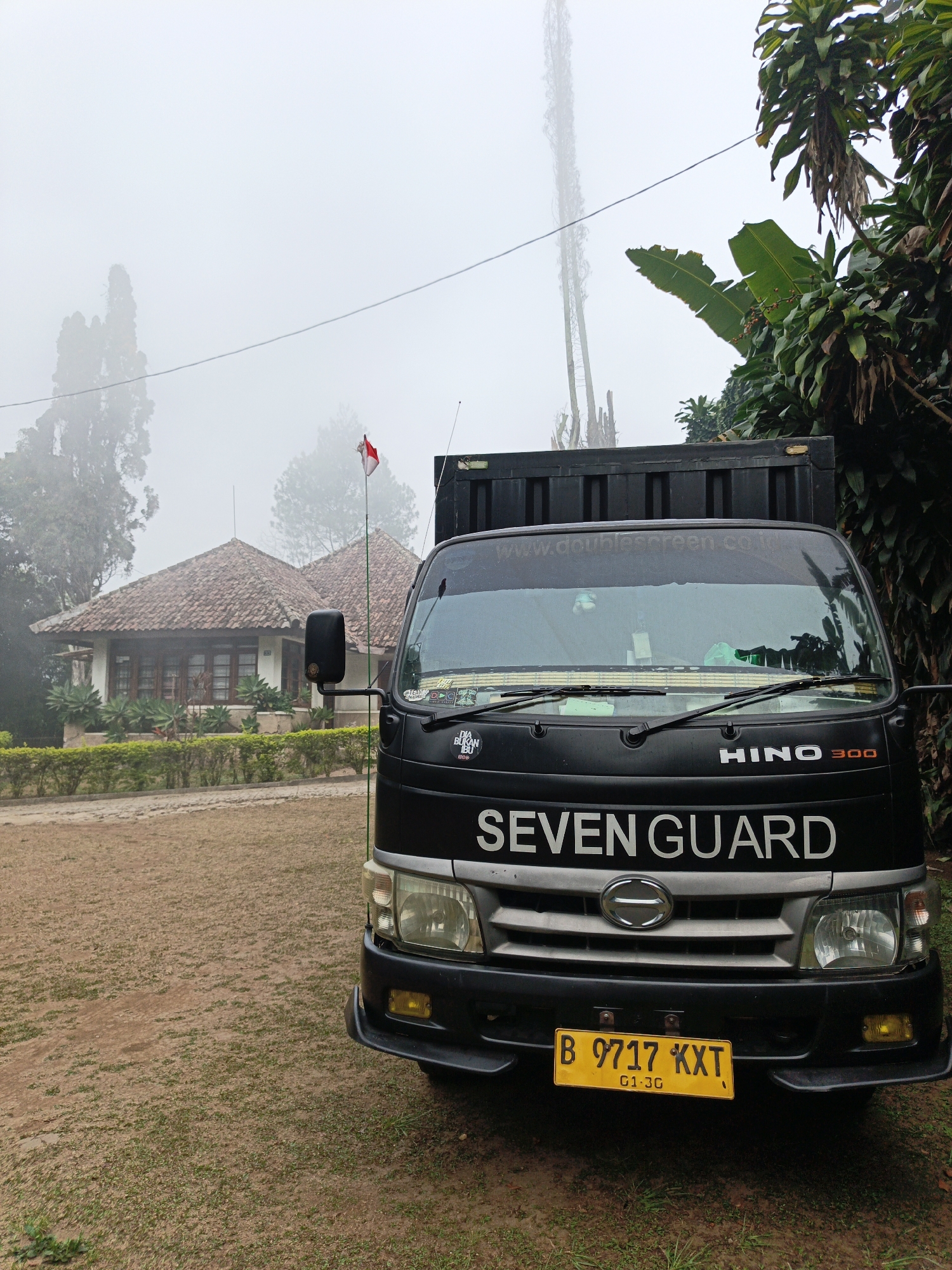 A black Hino 300 truck is parked on a dirt and grass lot in front of a single-story house with a red tile roof. The truck has "SEVEN GUARD" written in white on the front. The license plate is yellow with black lettering, reading "B 9717 KXT G1 3G". A small Indonesian flag is flying from a pole to the left of the truck. The scene is shrouded in fog, obscuring the background and creating a muted, overcast atmosphere. The house appears to be a dwelling, with manicured hedges and plants in front. Lush green trees and foliage surround the property. The overall impression is one of a quiet, perhaps rural, setting in Cibogo, Indonesia, characterized by the foggy weather.