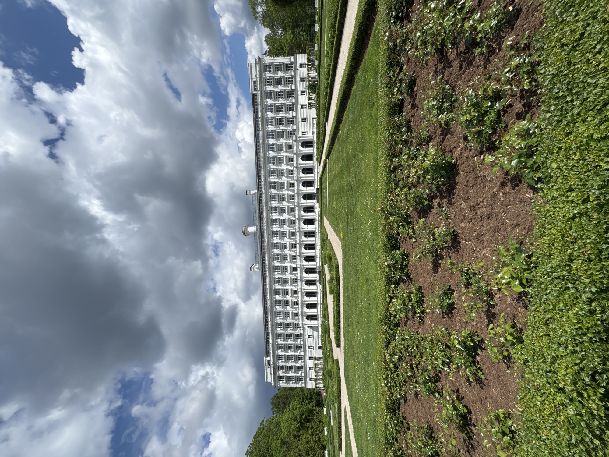 A low-angle, wide shot captures the grand facade of a white, multi-story building with a symmetrical design, reminiscent of a hotel or resort, situated in Jūrmala, Latvia. The building features numerous arched windows on its lower level and rectangular windows on the upper levels, all with white frames. A white balustrade runs along the roofline, and decorative chimneys are visible.
In front of the building is a meticulously maintained garden. The foreground features a bed of newly planted rose bushes, with rich brown soil and mulch surrounding them. Beyond this, a carpet of lush green grass is interrupted by manicured hedges and winding gravel paths, creating geometric patterns across the landscape. Further back, more manicured lawns and hedgerows lead towards the building. To the left of the building, dense green trees form a natural backdrop.
The sky above is dynamic, with a mix of dramatic, dark gray storm clouds and patches of bright blue sky, indicating a day with changing weather conditions. The overall scene conveys a sense of historical grandeur and natural beauty. No people or animals are visible in the image.