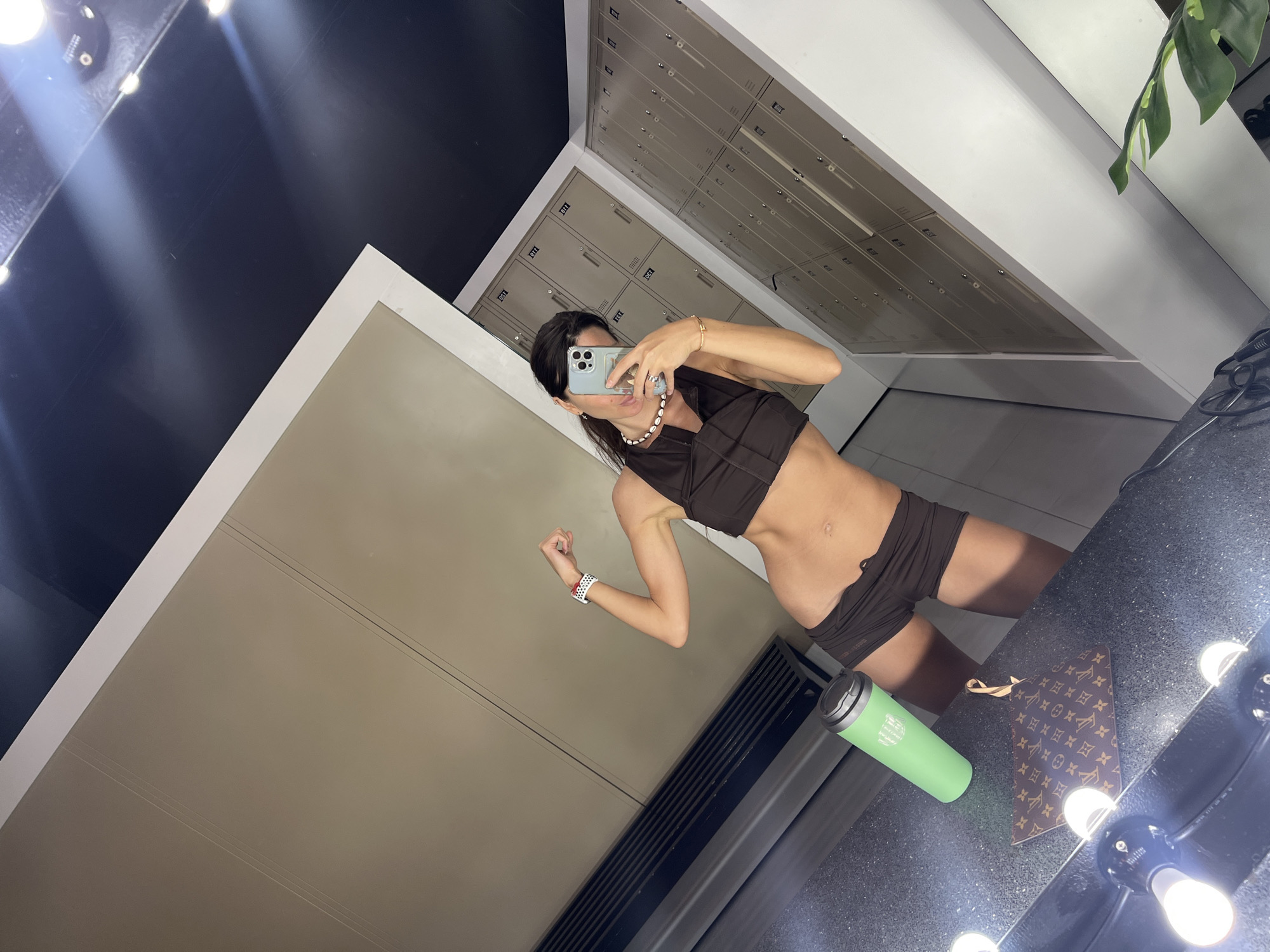 A woman is taking a mirror selfie in a locker room. She is wearing a brown sports bra and matching shorts. She is flexing her bicep and smiling. In the foreground, there is a green water bottle and a brown designer clutch. The background shows lockers and a bench. The lighting in the room is bright, with some lights visible in the mirror. The overall scene is clean and well-lit.