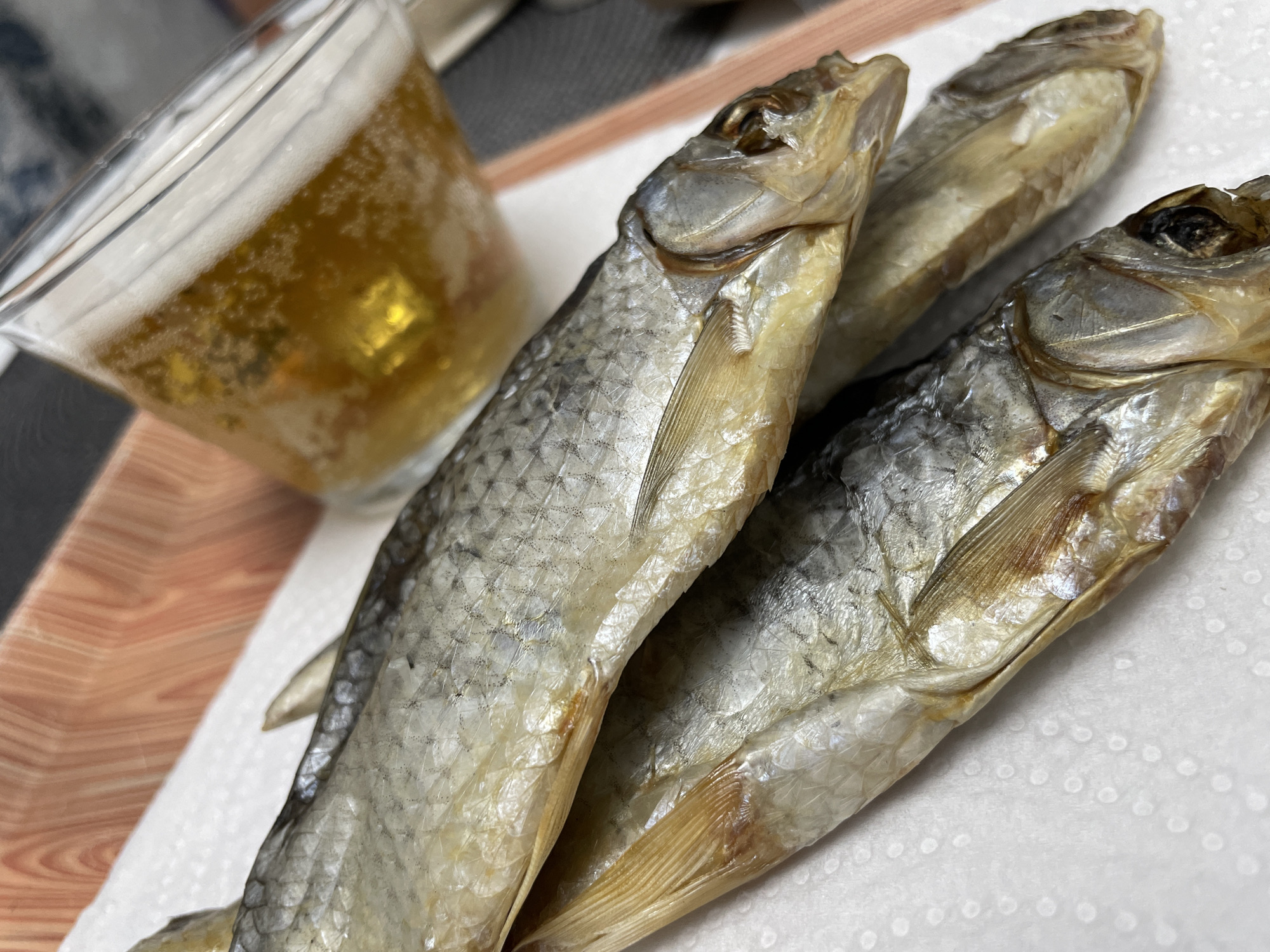 The image presents a close-up still life featuring three dried whole fish positioned alongside a glass of beer. The items are arranged on a white paper towel, which rests on a light brown surface with a distinct wood grain pattern, likely a tray or table.
The three dried fish are the primary subjects, arranged slightly overlapping each other. They appear to be small to medium-sized, with a characteristic dried and somewhat shriveled texture. Their scales exhibit a blend of silver-gray on the back and a lighter, yellowish-white on the belly. The heads are intact, showing visible, darkened eyes and gill covers. The fins are also distinguishable, pressed close to their bodies.
To the left of the fish, a clear glass holds a golden-yellow liquid topped with a frothy white head, indicative of beer. Numerous small bubbles are visible within the beverage, suggesting its effervescence. The glass is slightly out of focus, placing emphasis on the fish.
The white paper towel beneath the fish and glass has a subtle patterned texture. The underlying wooden surface provides a warm, natural background element.
The scene depicts a common culinary pairing, especially in Eastern European culture, where dried fish (such as vobla or taranka) is a popular snack often consumed with beer. Given the location context of Izhevsk, Russia, this arrangement strongly aligns with local traditions. There are no people visible, and the image captures a moment of readiness for consumption rather than an active event. No text is visible in the media file. The lighting appears even, suggesting an indoor setting.