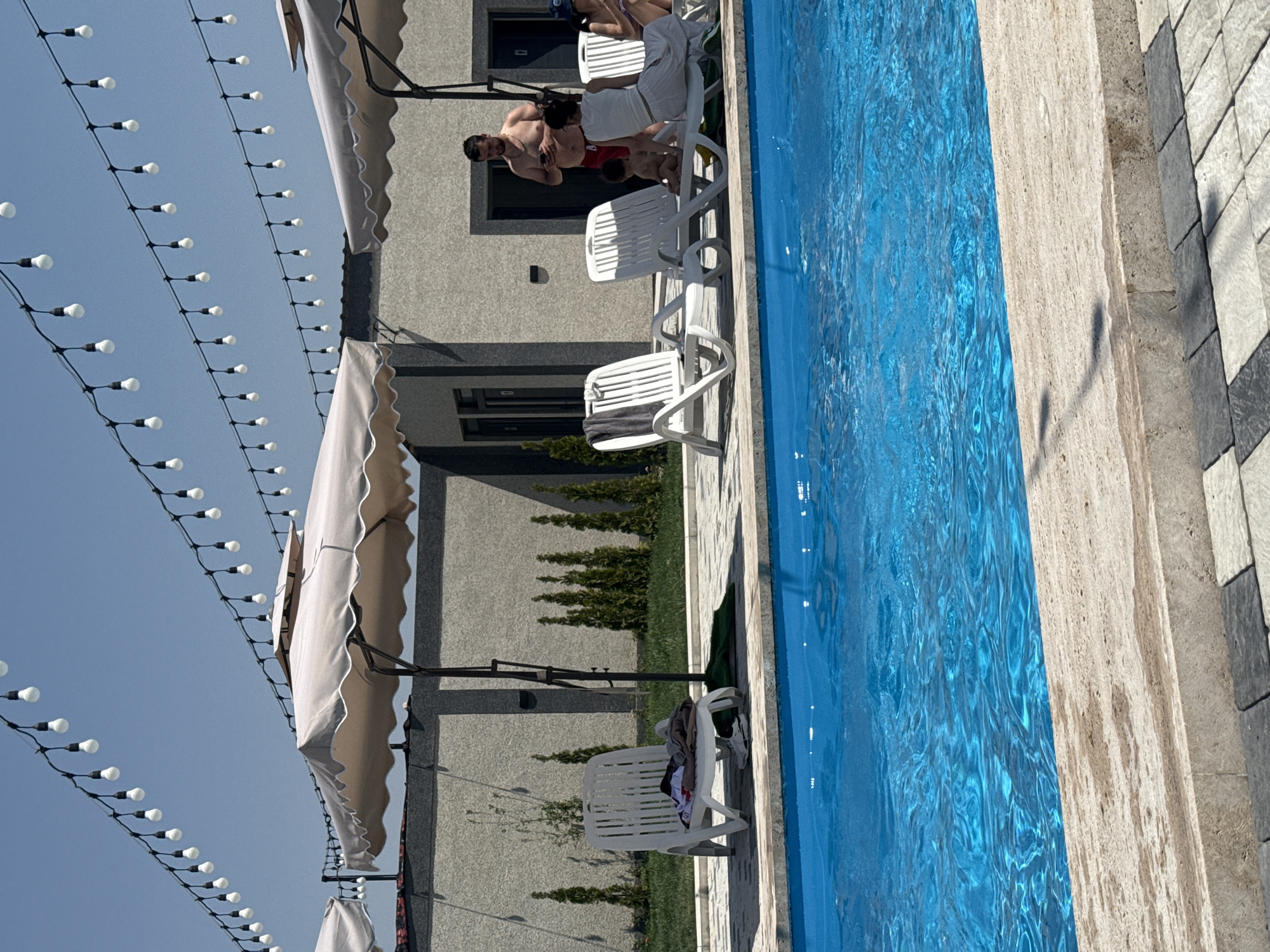 The image depicts a sunny day at a poolside establishment in Sasunik, Armenia. In the foreground, a bright blue swimming pool occupies the lower half of the frame, its surface rippling gently. The pool's edge is lined with light-colored stone coping, transitioning to a patterned paved area.
Beyond the pool, several white plastic lounge chairs are visible, some occupied by people. A man in red shorts stands near a child sitting on a lounge chair. A woman in a white top and skirt is also seated on a lounge chair, and another woman is partially visible in the background. Clothing is draped over one of the empty lounge chairs.
Large beige umbrellas provide shade over some of the seating areas. In the background, a modern building with a gray facade and dark-trimmed windows stands behind a neatly manicured lawn with rows of tall, slender evergreen trees. Overhead, strings of white lights are strung across the scene, suggesting it might be an area set up for evening enjoyment as well. The sky is a clear, bright blue.