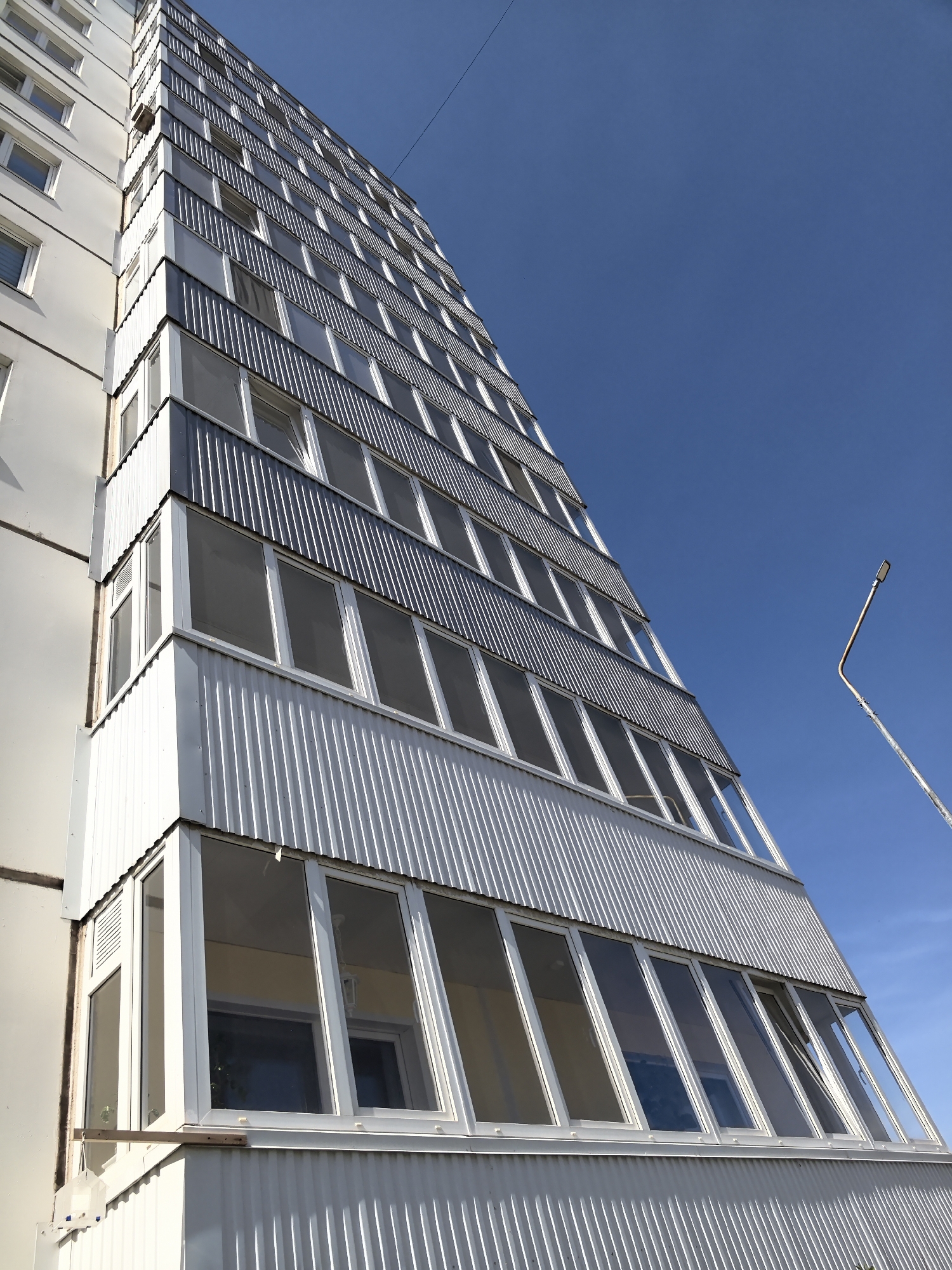 A low-angle shot shows a tall residential building in Фролы, Russia, against a clear blue sky. The building has a unique architectural design with balconies that protrude diagonally outward. The balconies are enclosed with corrugated metal panels and windows, giving the facade a ridged appearance. A street lamp is partially visible on the right side of the frame, adding to the urban context. The building's exterior is predominantly white and gray, contrasting with the blue sky.