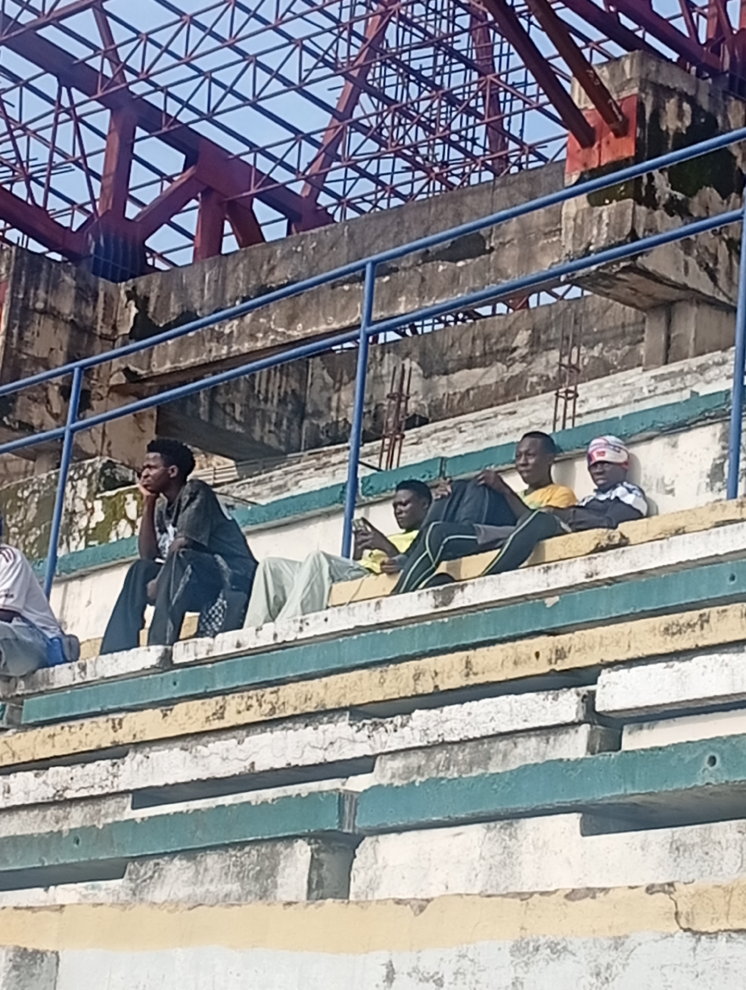 In New Stadium, Nigeria, a group of young men are seated on tiered concrete bleachers, likely at a sports venue. The bleachers are painted in alternating bands of yellow, white, and teal, showing signs of wear and tear with chipped paint and weathered concrete. A blue metal railing runs along the top of the seating area. In the background, a complex red metal structure, possibly part of a stadium roof or support system, is visible against a clear blue sky. The men are casually dressed and appear to be observing something off-camera. One man in the foreground is leaning back with his hand on his chin, looking contemplative. Others are seated next to him, some looking forward, one with a head covering. The overall scene suggests a relaxed atmosphere in an outdoor public space.