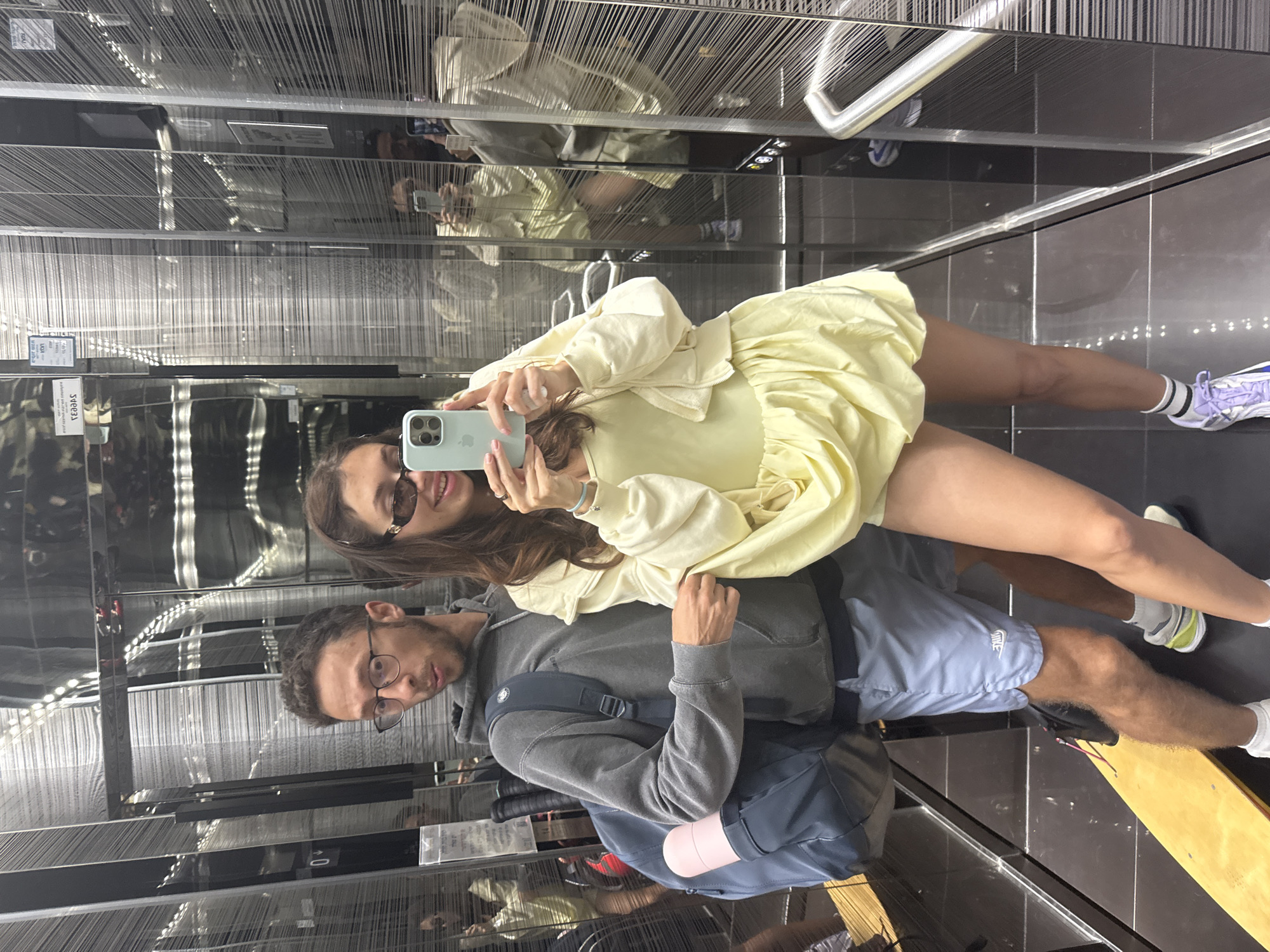 Two individuals pose for a selfie in an elevator featuring mirrored walls with vertical decorative strings. The woman, on the upper right, wears a yellow dress and holds a light blue smartphone. She is wearing sunglasses. The man, slightly behind and to the left of the woman, has short hair, wears glasses, and is dressed in a gray hooded sweatshirt, blue shorts, and carries a dark blue backpack with a pink water bottle attached. Reflections in the mirrored elevator walls create a repetitive effect, showing multiple iterations of the couple and the elevator interior. The elevator floor consists of dark grey tiles. The presence of the mirrored elevator interior indicates a modern building or establishment, possibly located in Tel-Aviv, Israel.