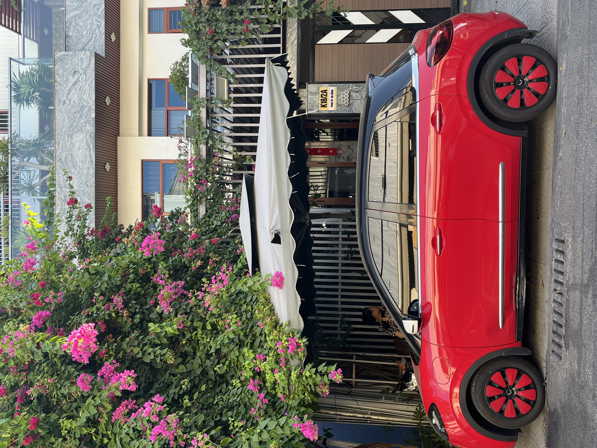 The image shows a red and black car parked on a street in Đà Nẵng, Vietnam. A bougainvillea bush with pink flowers is on the left side of the frame. Behind the bush is a building with a white awning. The building's address, K18/2A Phan Tử - Đà Nẵng, is visible on a sign. The building has a white exterior with wooden details. The street is concrete and appears to be well-maintained. There is a person inside the building that can be seen reflected on the windows of the car.