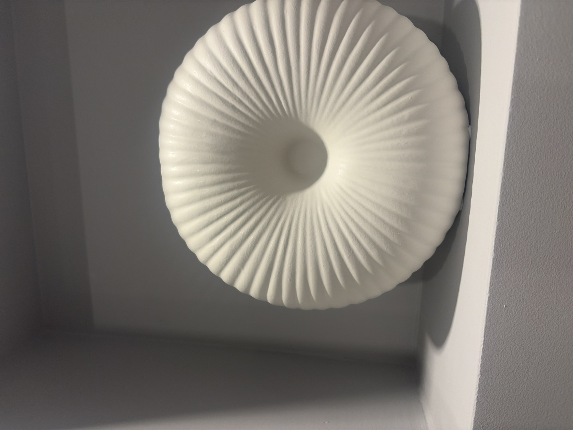 A white, disc-shaped object with radiating ridges and a hollow center rests on a white shelf in a grey, recessed area, likely a display niche or shelf. The object appears to be made of ceramic or a similar matte material. The lighting suggests an indoor environment. This image was taken in Podgumer, Bulgaria.