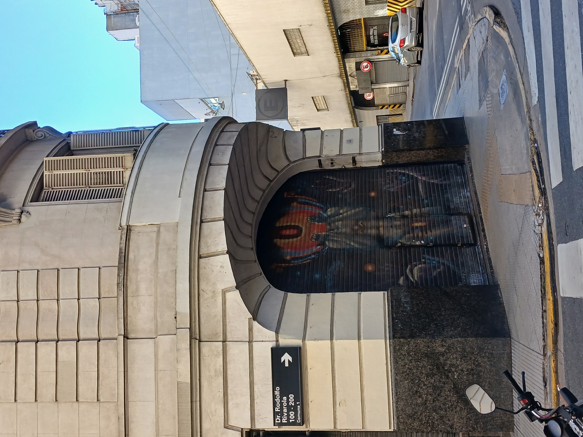 The image shows a street view in Buenos Aires, Argentina, specifically featuring the entrance of a building with an arched mural. The building is constructed with light-colored stone or concrete blocks, and the mural displays a dark-toned abstract painting behind a wrought iron gate. A street sign points to Dr. Rodolfo Rivarola 100-200, Comuna 1. The street is paved, with a motorcycle partially visible in the foreground. In the background, taller buildings can be seen, along with traffic signs and a car parked on the side of the road. The sky is a light blue color, indicating daytime.