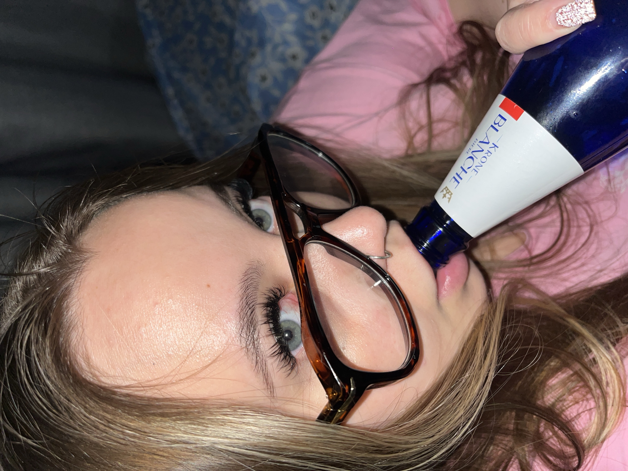 A woman with light blonde hair is tilted sideways, wearing glasses, drinking from a blue and white Krone Blanche beer bottle. She has a pink shirt on and there is a blue flowered background. The location is unknown and there is no indication of Krasnodar, Russia.