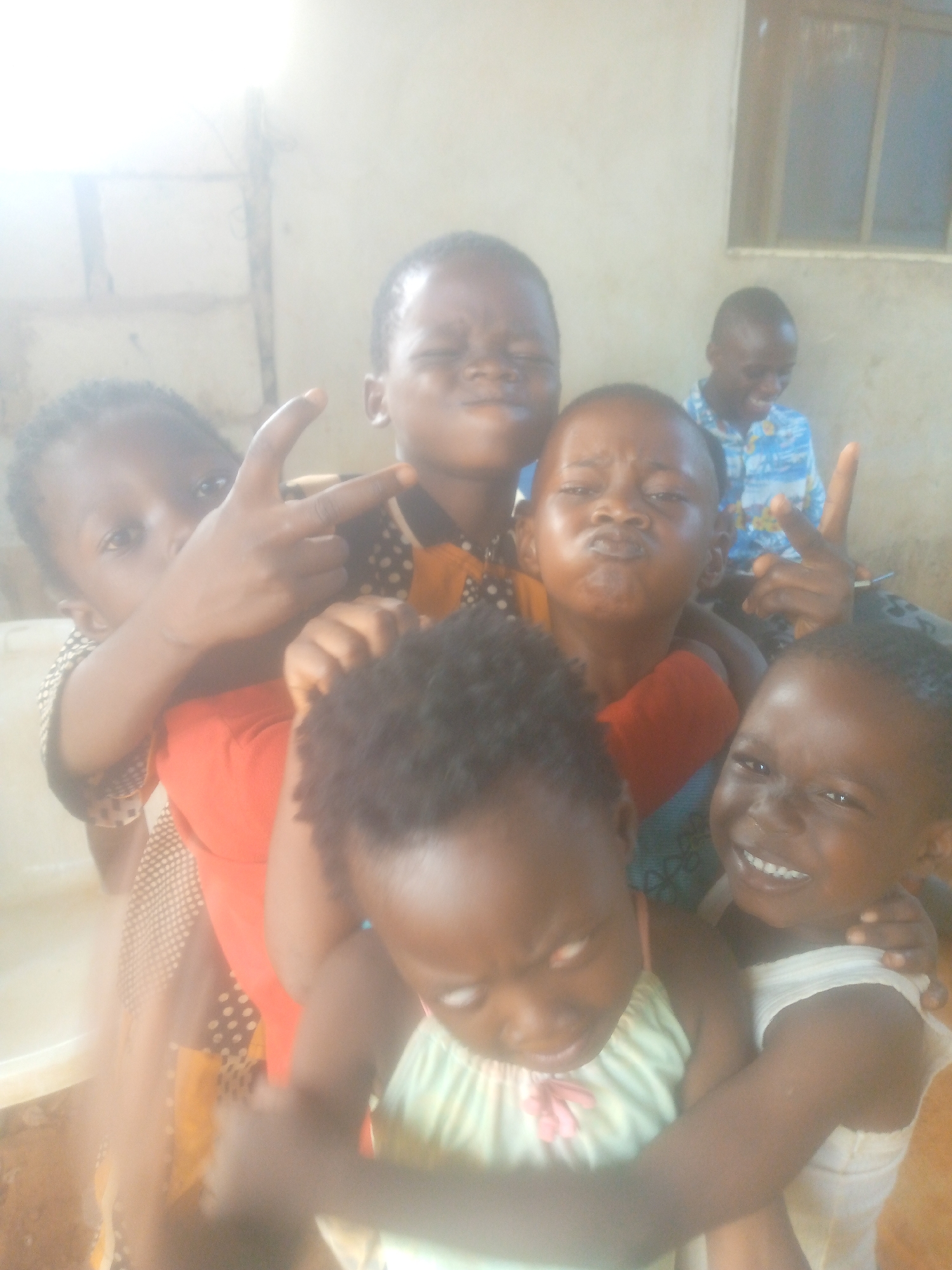 A group of six children and one man are indoors. In the foreground, five children are gathered close together, with some making hand gestures. The children have dark skin and are dressed in various casual clothing. In the background, a man is seated, wearing a blue patterned shirt and also making a hand gesture. A window with bars is visible behind him. The lighting in the room appears bright, but there's also some glare. The location is in the city of Unknown, Unknown.