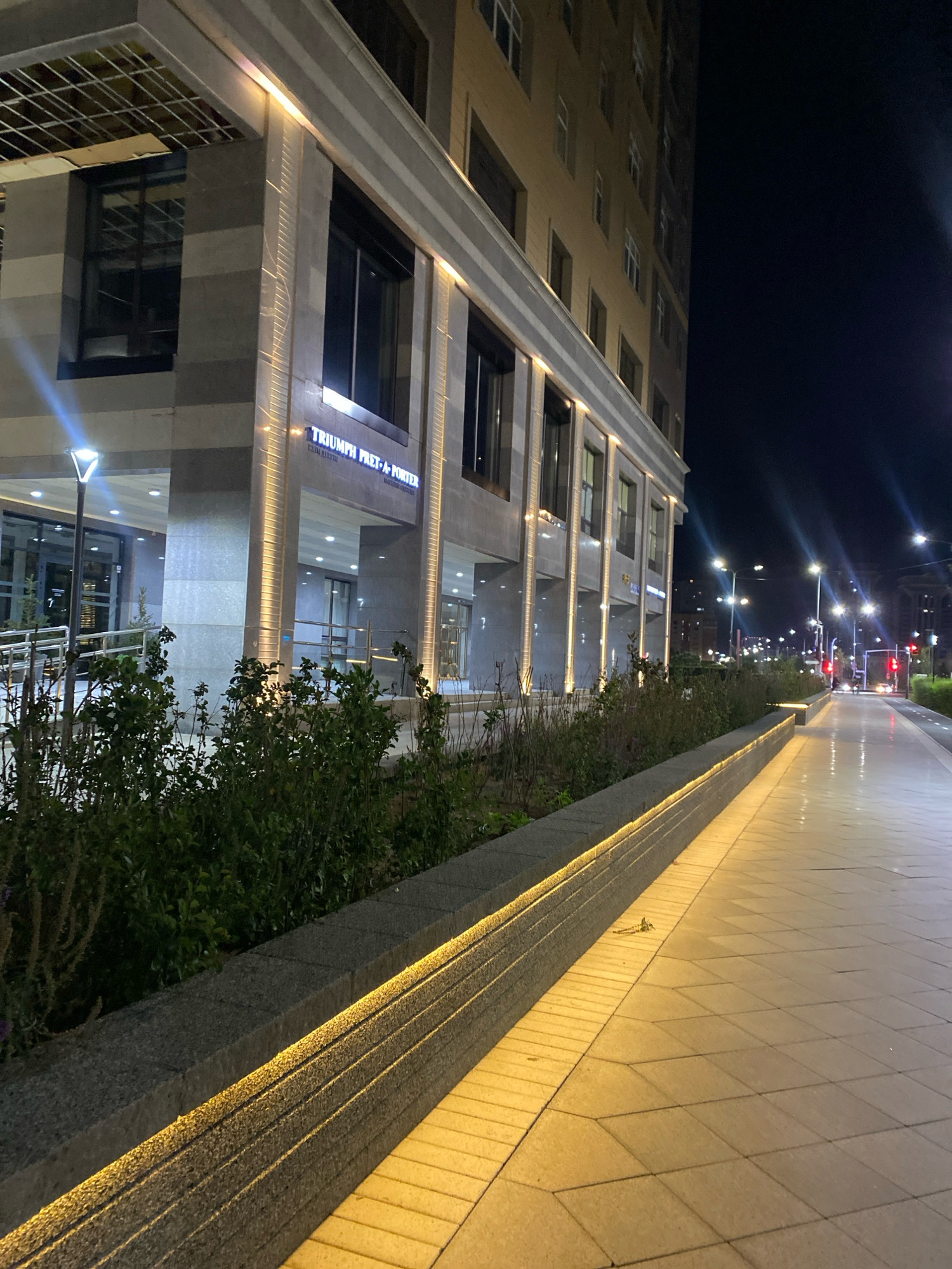 The image captures a nighttime view of a modern building facade and an adjacent pedestrian walkway in Astana, Kazakhstan. The building, featuring a contemporary architectural style with horizontal banding of stone and vertical illuminated columns, displays a sign that reads "TRIUMPH PRET-A-PORTER." To the left of the building entrance, a tall, modern lamppost illuminates the scene. In the foreground, a well-maintained planter box filled with lush green shrubs runs along the edge of the walkway. The walkway itself is paved with large, light-colored tiles and is illuminated by streetlights, casting a warm glow and creating distinct shadows. In the distance, the street is visible with traffic lights, car headlights, and the silhouettes of other buildings against the dark night sky, indicating an urban environment. The overall impression is one of a well-lit, well-maintained urban public space at night.