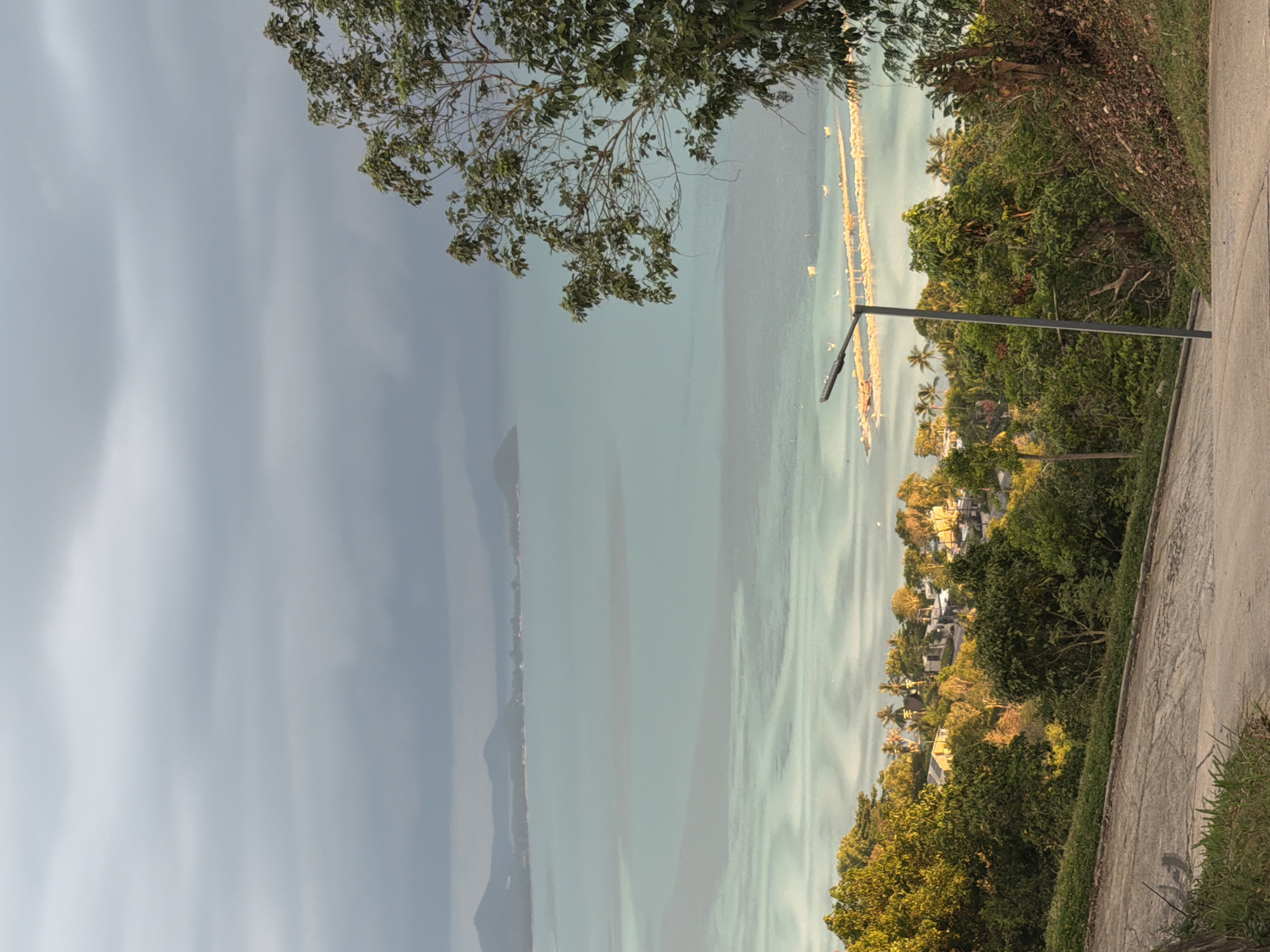 The image depicts a scenic view of the coast, likely in or near Baan Thong Po, Thailand, under an overcast sky. In the foreground, a paved pathway curves downwards, bordered by grass and trees on the left and a dirt embankment on the right. A tall, dark gray lamppost stands near the path, casting a shadow. Beyond the immediate foreground, a dense green forest covers a hillside, interspersed with buildings and palm trees, suggesting a residential area.
The middle ground features the tranquil sea, characterized by its light turquoise hue and visible underwater sandbars or shallow areas that create swirling patterns. In the distance, a long, low structure, possibly a pier or a breakwater, extends into the water, lined with smaller structures that appear to be fishing boats or buoys. Further out, a chain of islands with hazy mountain silhouettes forms the horizon. The overall atmosphere is serene and natural. There are no people visible in the image.