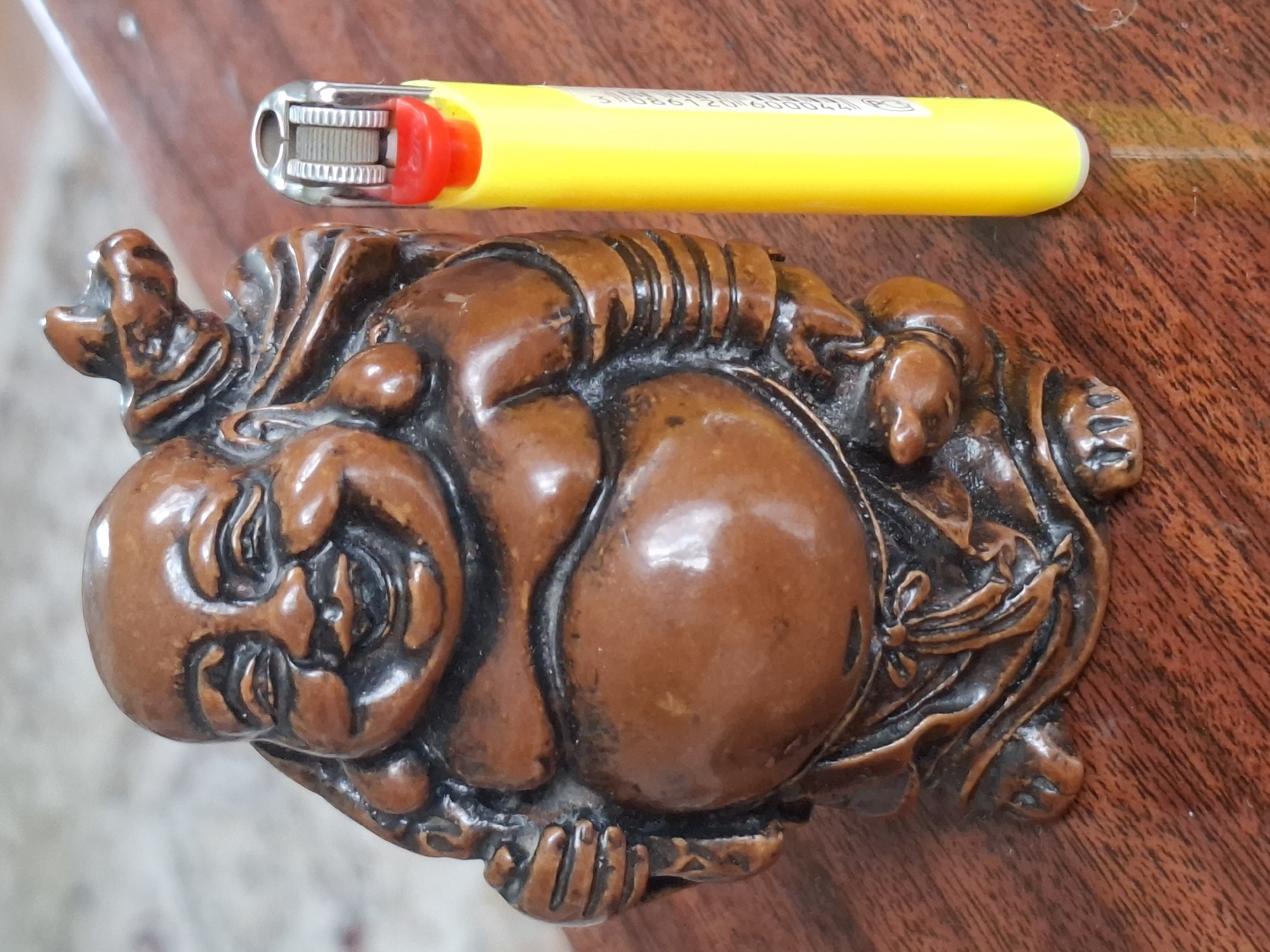 This media file features a close-up, overhead shot of a carved wooden figurine and a yellow lighter, both resting on a polished wooden surface. The figurine appears to be a depiction of a laughing Buddha, characterized by its rounded belly, smiling face, and intricately carved details of clothing and possibly other figures or ornaments surrounding it. The wood is a rich brown color with visible grain and a glossy finish. The lighter is bright yellow with a red igniter. Text is visible on the yellow lighter, appearing to be a series of numbers and letters, possibly a serial number or branding. The lighting suggests an indoor setting, likely during daylight hours, with the light source creating subtle reflections on the smooth surfaces of the objects and the table. The background is blurred, but the wooden texture of the table is clearly discernible. The scene is static, with no apparent activity taking place beyond the placement of the objects. The overall impression is one of still life, possibly showcasing a decorative item or personal belonging. The location context of Almaty, Kazakhstan, provides a geographical setting but no specific visual cues within the image directly indicate this location.