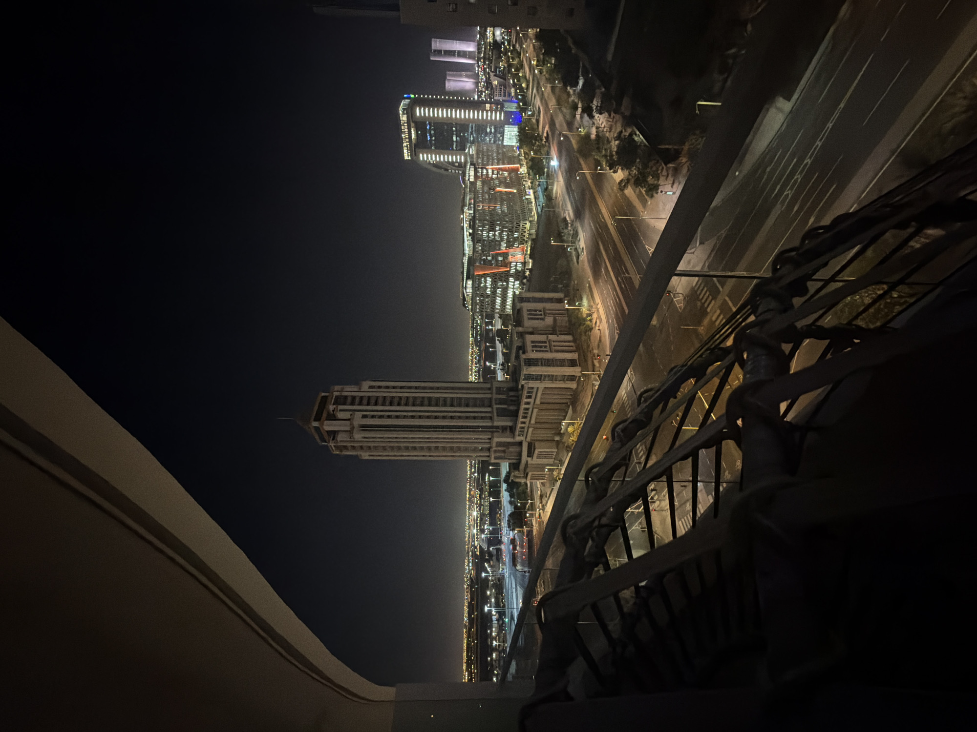 This image captures a wide, elevated view of an urban cityscape at night, likely from a balcony or high-rise window. In the immediate foreground, the dark, multi-bar metal railing of a balcony is visible across the lower third, with a light-colored, curved architectural element forming an overhead arch in the upper left.
The central mid-ground is dominated by a prominent, tall, light-colored building with intricate, multi-tiered architecture and a slender spire, identified as the Abu Dhabi Courts Complex / Judicial Department. This building has a wider, multi-story base that tapers into the main tower.
In front of this complex, a multi-lane urban road, Sheikh Rashid Bin Saeed Street, stretches across the mid-ground, illuminated by numerous streetlights. A large white and red bus is visible on the road, along with other less distinct vehicles. Pedestrian crossings and traffic signals are also visible on the street.
Further in the background, to the right, a cluster of modern, illuminated high-rise buildings on Al Maryah Island stands out against the dark sky. These include structures with distinct vertical strip lighting and geometric facades, characteristic of the Abu Dhabi Global Market (ADGM) complex.
The distant horizon is marked by a vast expanse of countless city lights, indicating a sprawling metropolitan area. The sky above is dark and clear, typical of a night scene. No discernible individuals or specific actions are visible, presenting a static depiction of a well-lit, quiet urban landscape at night in Abu Dhabi, United Arab Emirates.