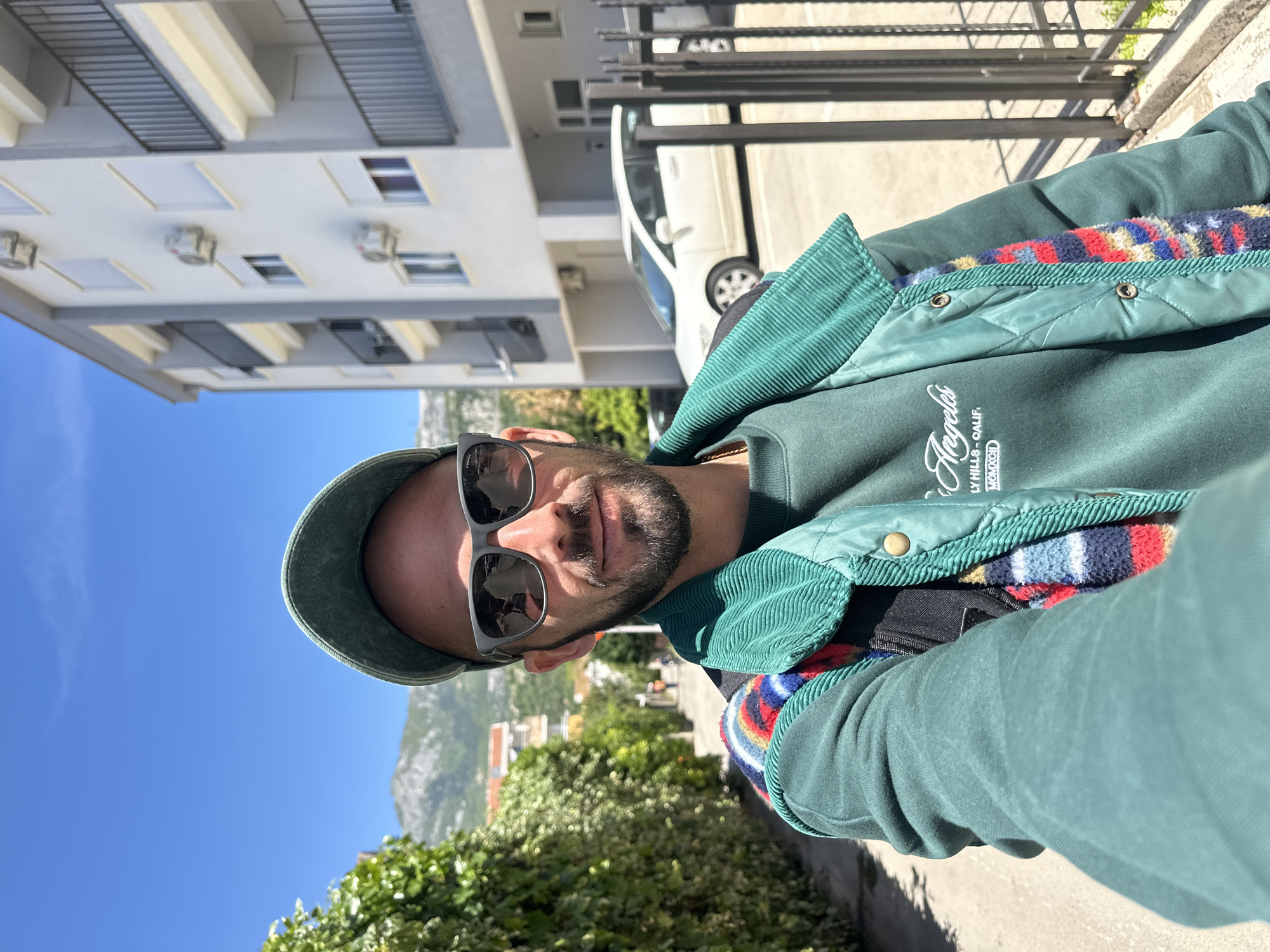 A man takes a selfie outdoors in Budva, Montenegro, under a clear, bright blue sky. The image is rotated 90 degrees clockwise. He is depicted from the chest up, wearing a dark green cap, dark sunglasses, and a dark green corduroy jacket with a quilted panel. A colorful, patterned lining is visible inside his jacket. He wears a dark green sweatshirt underneath, which has white text: "Angeles" in a script font, followed by "BEVERLY HILLS - CALIF." and "MCMXCII". A black backpack strap is visible on his shoulder.
The man has a beard and a neutral to slightly smiling expression, partially hidden by his sunglasses. The strong sunlight casts shadows, indicating a sunny mid-day or afternoon.
In the background, behind green foliage, are modern, light-colored multi-story buildings with balconies, suggesting a residential or urban street. Cars are also visible on what appears to be a street or parking area. In the far distance, rugged, rocky mountains rise prominently, characteristic of the Montenegrin landscape. The setting combines urban elements with striking natural scenery.