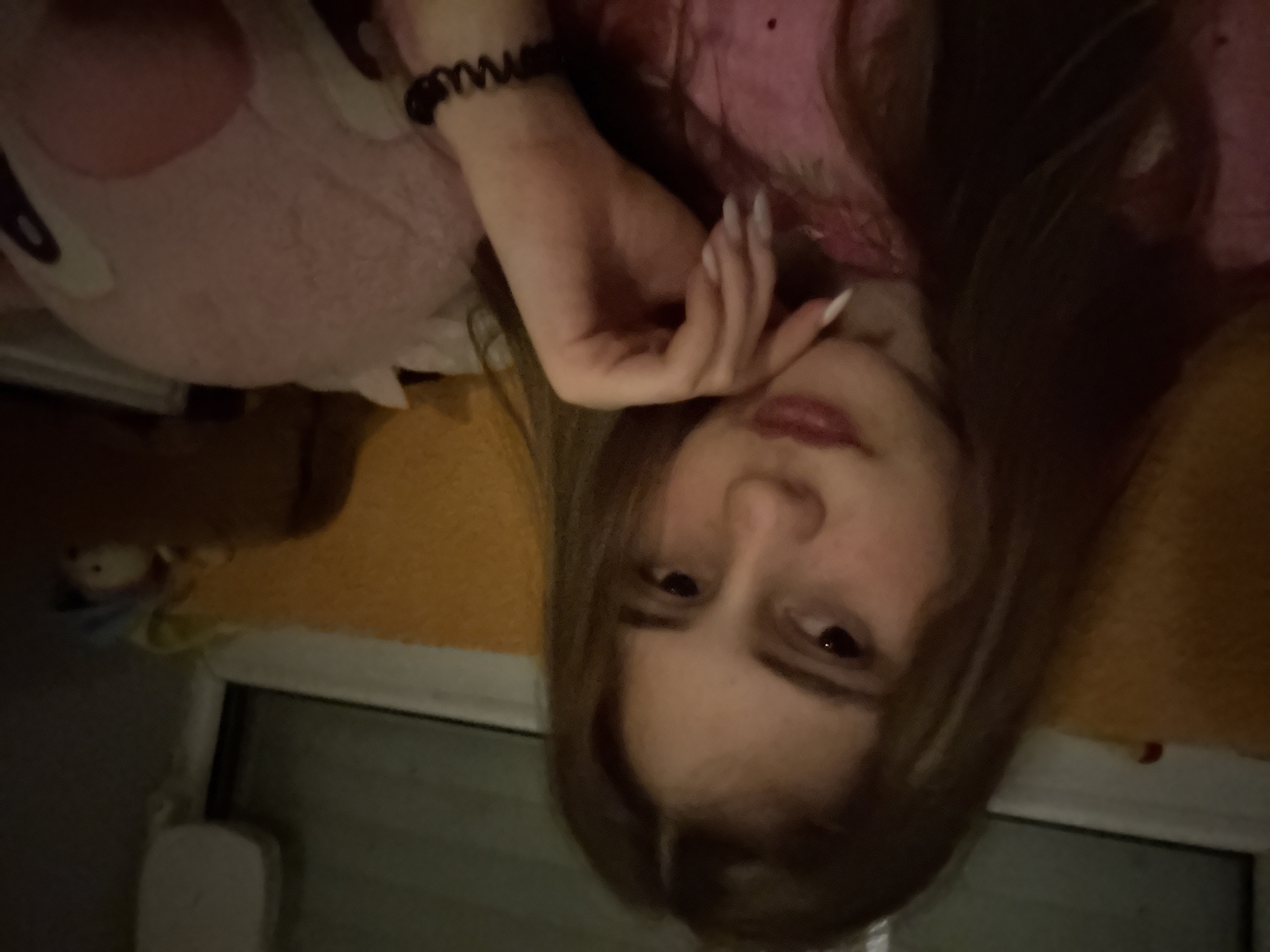 A young woman with long brown hair is lying down. She is wearing a pink top and a black spiral hair tie on her wrist. Her left hand is resting near her face, showing long, white fingernails. A pink plush toy is next to her. The scene appears to be indoors, possibly a bedroom, with a window visible in the background. The location is not identifiable as Panagyurishte, Bulgaria from the visual information provided.