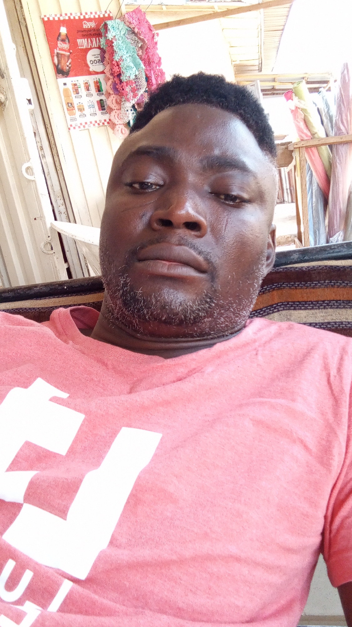 A medium shot shows a dark-skinned man wearing a pink T-shirt with a large white letter on the front, presumably part of a word. The man has a close-cut, dark hairstyle and a short, graying beard. He appears to be indoors, possibly in a shop or small business, as evidenced by a Coca-Cola advertisement and other posters on a wall behind him. Assorted textiles and a simple wooden structure can also be seen in the background. This suggests a scene in Sabon Tasha, Nigeria, a bustling commercial area with numerous shops and market stalls.