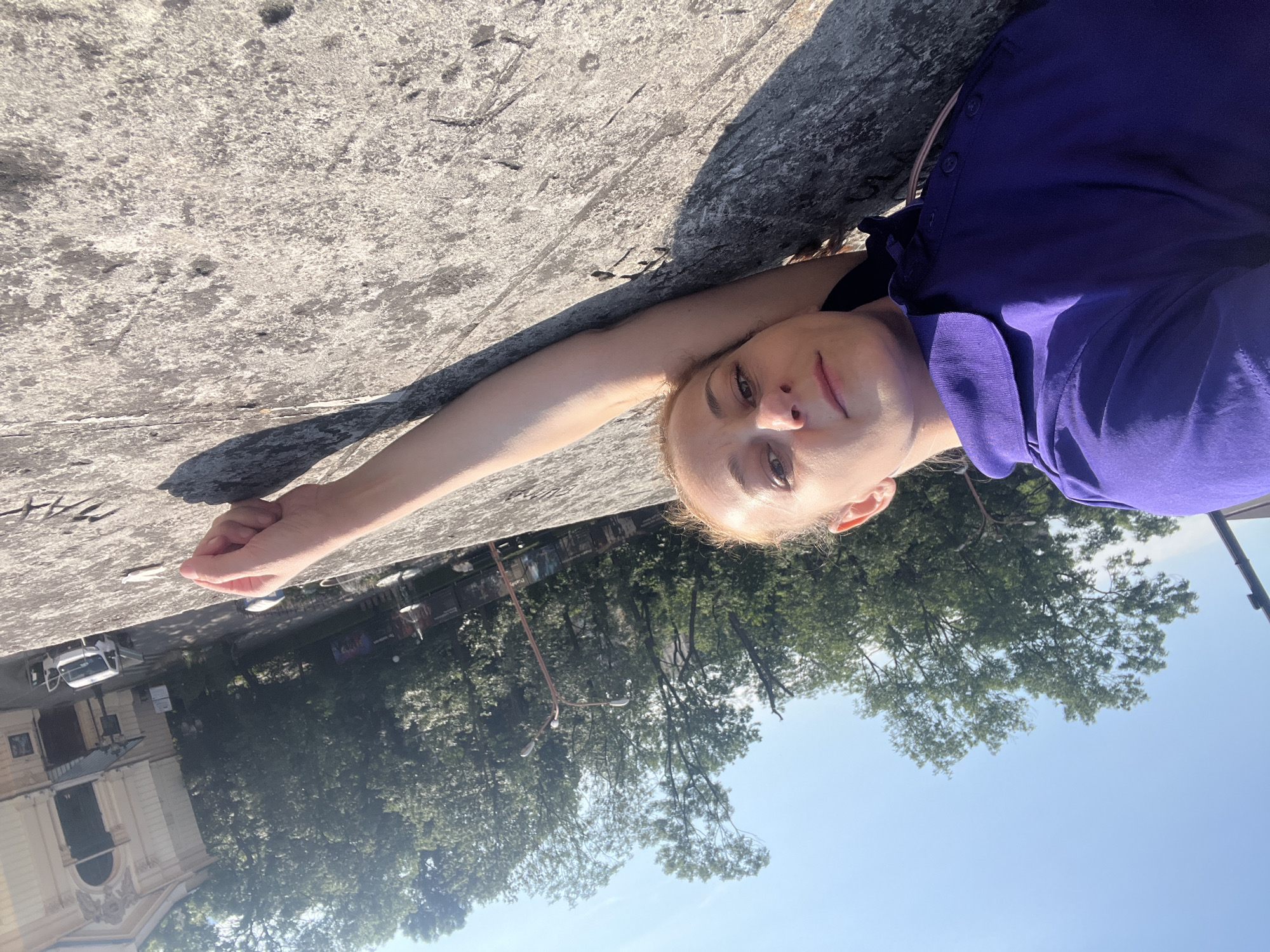A blonde woman lies on a stone surface under a clear blue sky, her head resting on her left arm which is extended to the right. She is wearing a purple collared polo shirt. Her right arm is visible extended in front of her. Trees and a light-colored building with an arched entrance are in the background. A white pickup truck with its doors open is parked in front of the building. The surface she is lying on appears to be a wall or embankment, with some etched markings visible.