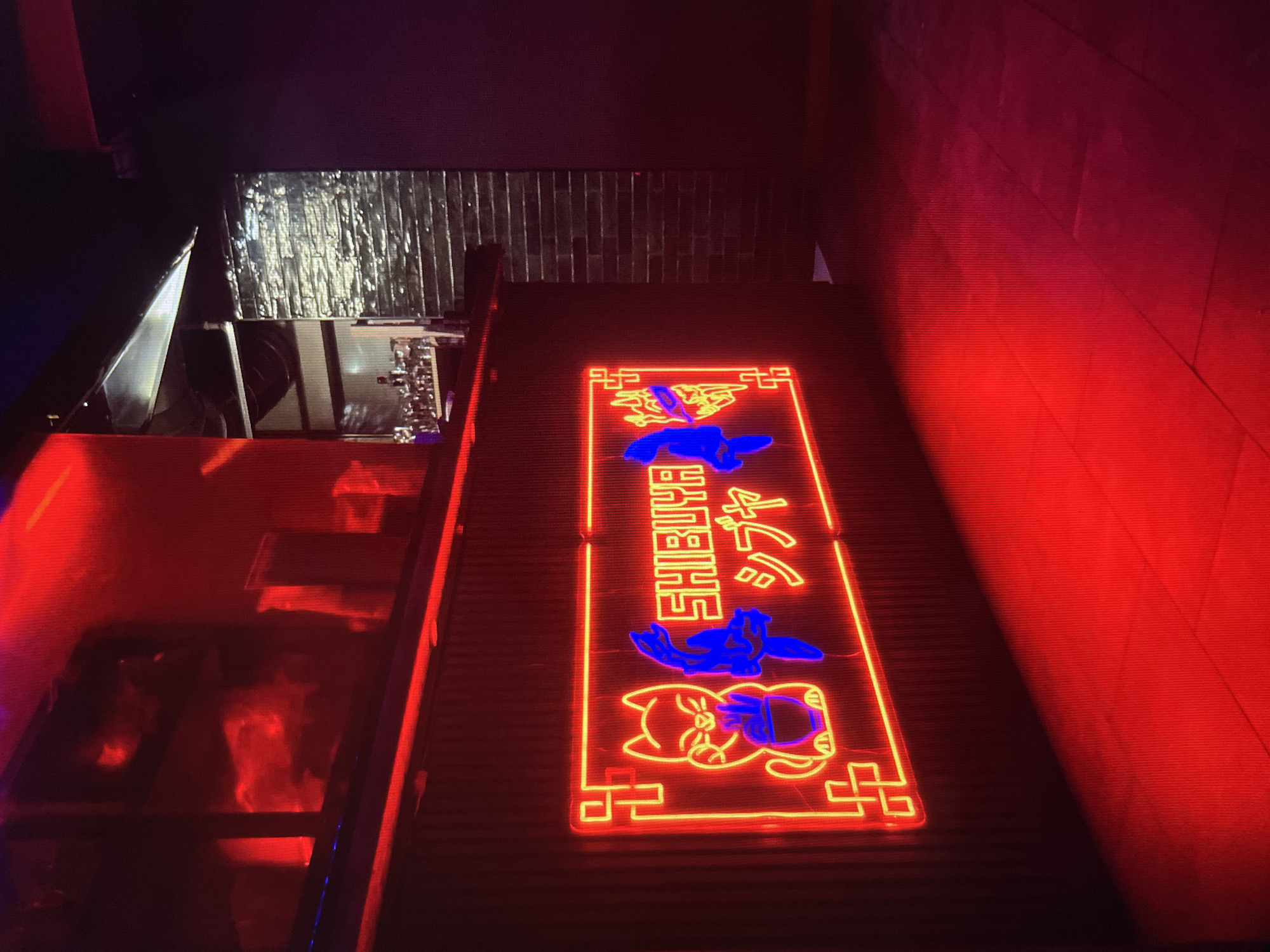 The image depicts a neon sign reading "SHIBUYA" and possibly a Japanese translation. The sign features a red border and yellow outlines of cartoon cat images. The text "SHIBUYA" is blue. The sign is mounted on a dark surface, likely a building exterior. The lighting is low with a strong red cast dominating the right side of the image. To the left of the sign is a reflective metallic surface above an area illuminated with red light, possibly a window or opening reflecting the red lighting. The location is Shibuya, Japan based on the sign.