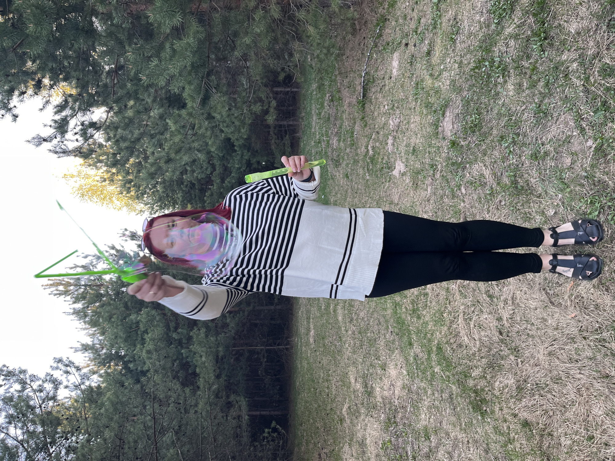 A person is outdoors, standing on a grassy area surrounded by trees, with the sun shining. The person has red hair and is wearing a black and white striped sweater and black leggings. The person is holding a green bubble wand in front of them and blowing bubbles. In their left hand is a second wand. The person is also wearing black sandals. The location may be in the city of Chaikovsky, Russia.