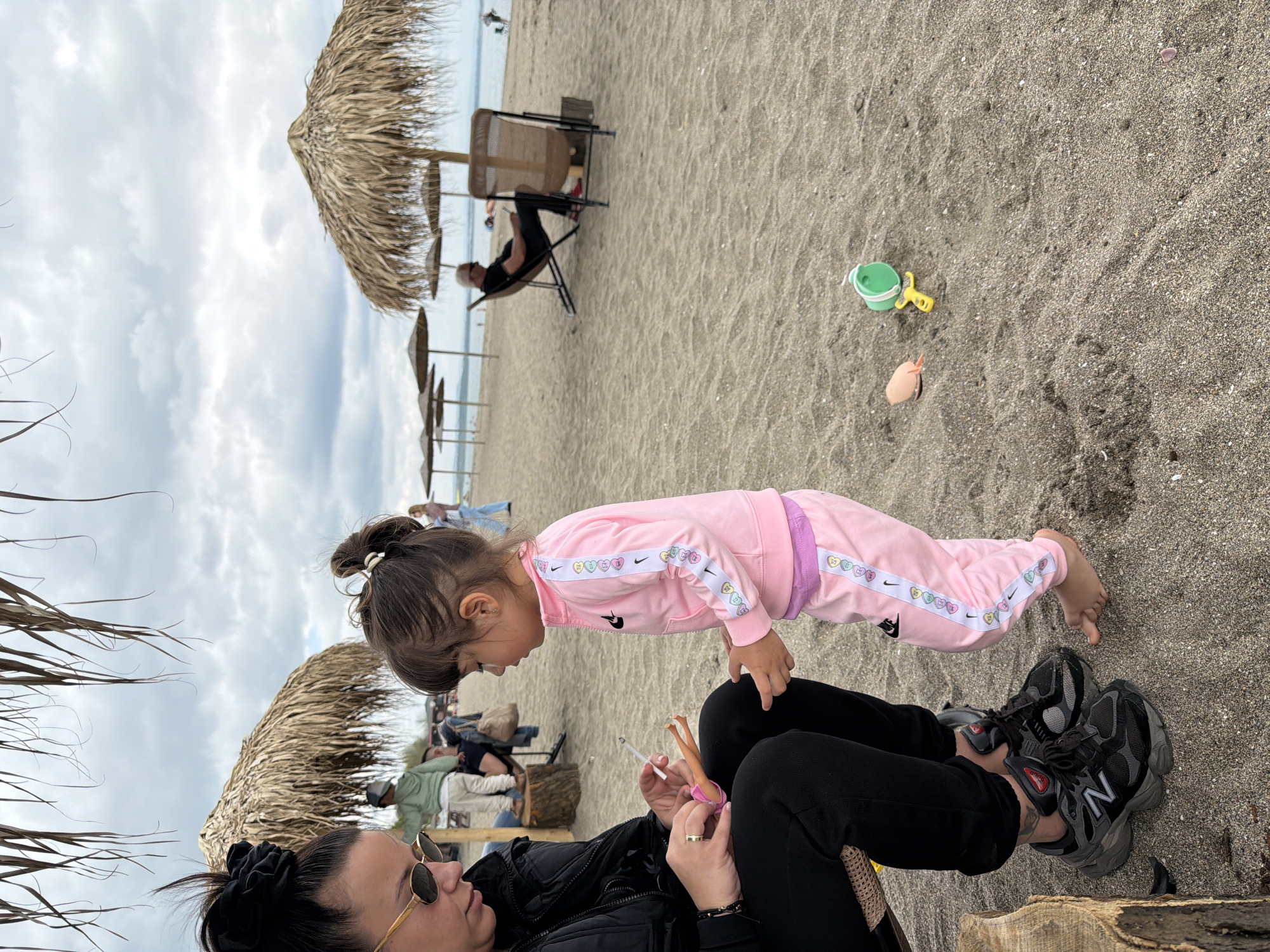 The image captures a scene at the beach. A child wearing a pink tracksuit is standing on a woman's lap. The woman is wearing black pants and a black jacket, with sunglasses. She is sitting on a piece of wood. A doll is in her hand. In the background, there is sand, several beach umbrellas, and other people. The sky is cloudy. A small bucket and shovel are on the sand.