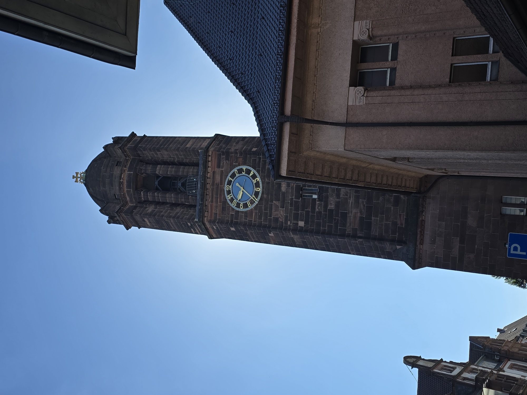 The image shows the tower of a church, likely the Luther Church in Worms, Germany, against a clear blue sky. The tower is made of dark brick and features a clock with a blue face and gold hands and Roman numerals. The sun is positioned behind the tower, creating a lens flare effect. A portion of the church's roof, covered in dark tiles, is visible in the foreground on the right, obscuring part of the tower. The architectural style suggests it is a historic building. In the background on the left, there are parts of other buildings with pitched roofs, also made of brick and featuring visible window frames. A blue parking sign with a white 'P' is visible near the bottom of the image. The scene is captured from a low angle, looking upwards.