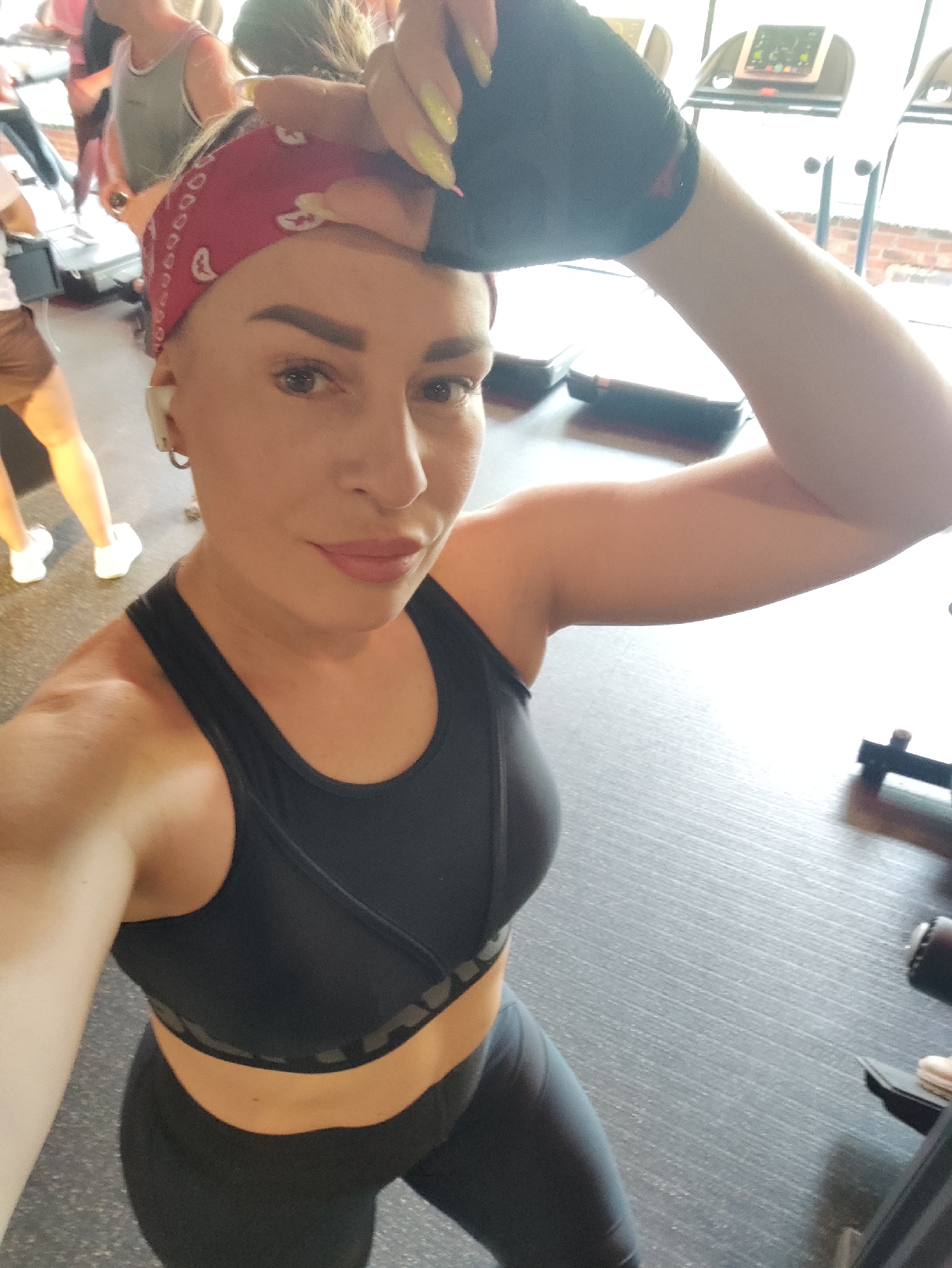 A woman with blonde hair tied back with a red bandana and wearing a black sports bra and black leggings is taking a selfie in a gym. She is wearing black workout gloves and appears to be wiping sweat from her forehead. In the background, there are treadmills and other gym equipment, as well as other people working out. The floor is a dark gray, textured surface. The image is taken from a high angle, looking down at the woman. The lighting is bright, suggesting indoor gym lighting. There are no specific landmarks or indicators that would place this scene in Almaty, Kazakhstan.