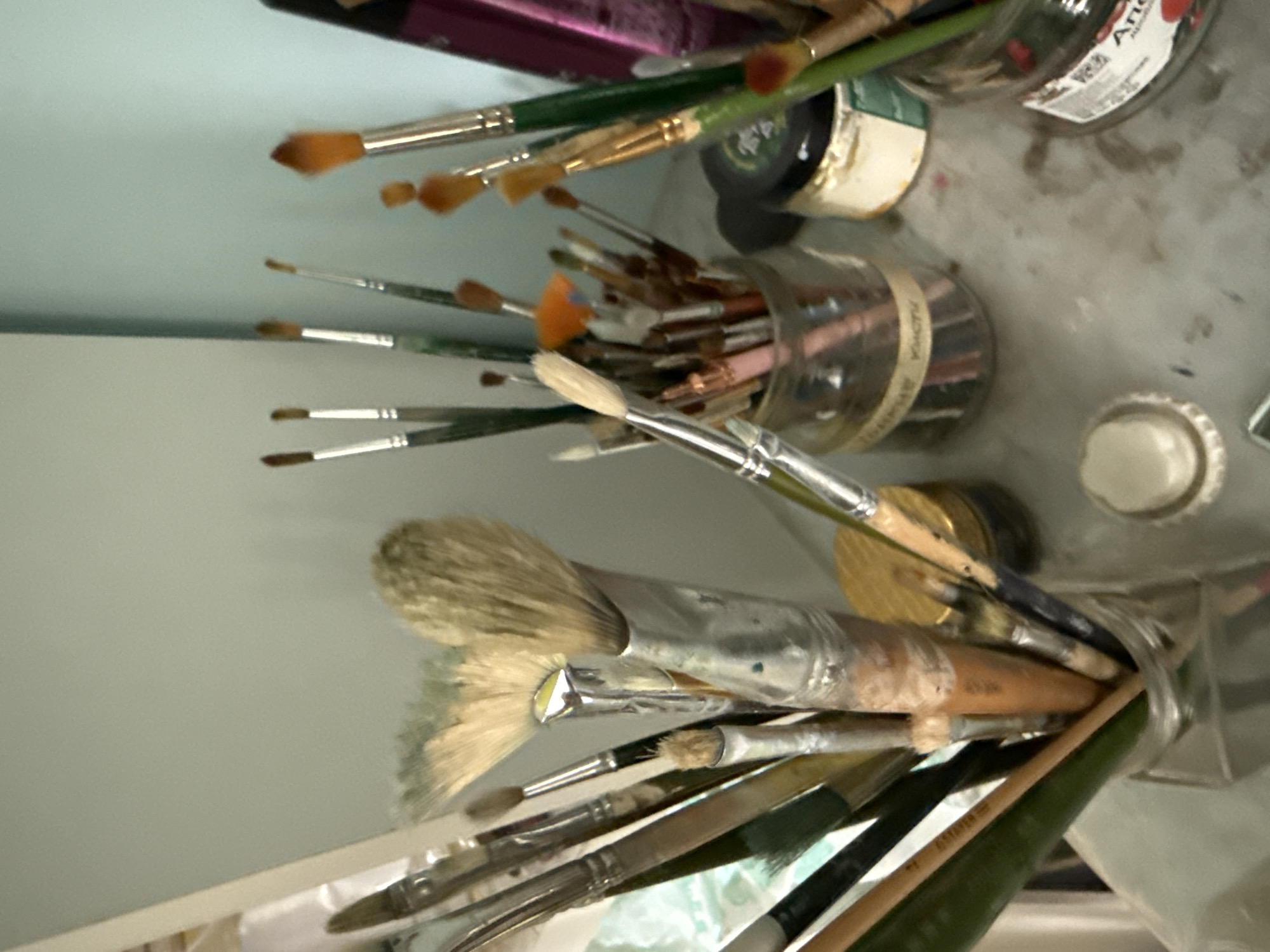 A still life composition of assorted paintbrushes in glass jars on a cluttered surface. The paintbrushes vary in size, bristle type, and handle color, with many showing signs of use. Some of the jars contain other art supplies, such as small containers of pigment or medium. The background is a light blue wall. The overall scene suggests an artist's workspace, potentially in Vladivostok, Russia, given the presence of some Cyrillic labeling on the jars, though this cannot be definitively confirmed without clearer views of the text. The lighting is soft and diffused, creating a slightly muted color palette.